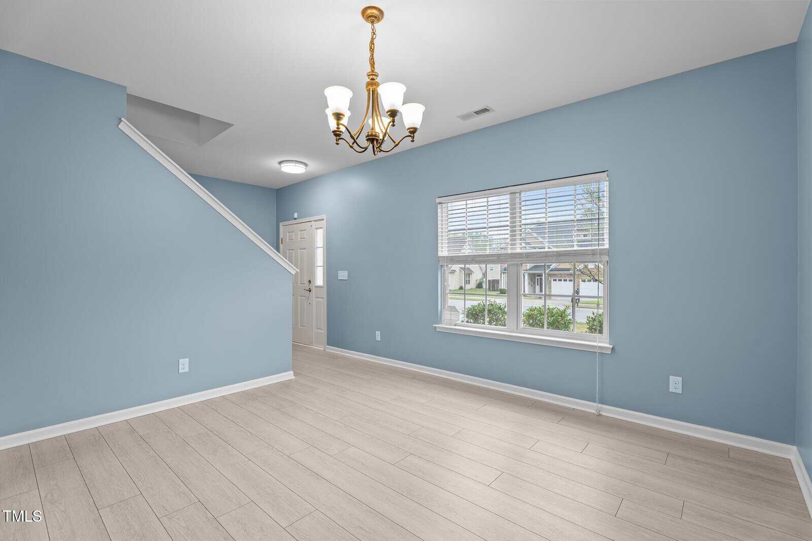 A room with light blue walls and white trim features a wooden floor, a window with blinds, a staircase with a white railing, and a hanging chandelier. There is a door beside the staircase. Natural light enters through the window.