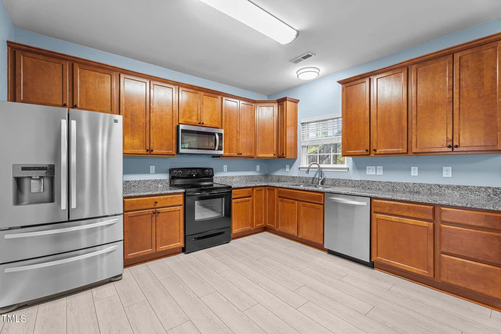 A modern kitchen with wooden cabinets, granite countertops, and stainless steel appliances including a refrigerator, dishwasher, and microwave. A window above the sink provides natural light. The floor is covered with beige tiles.