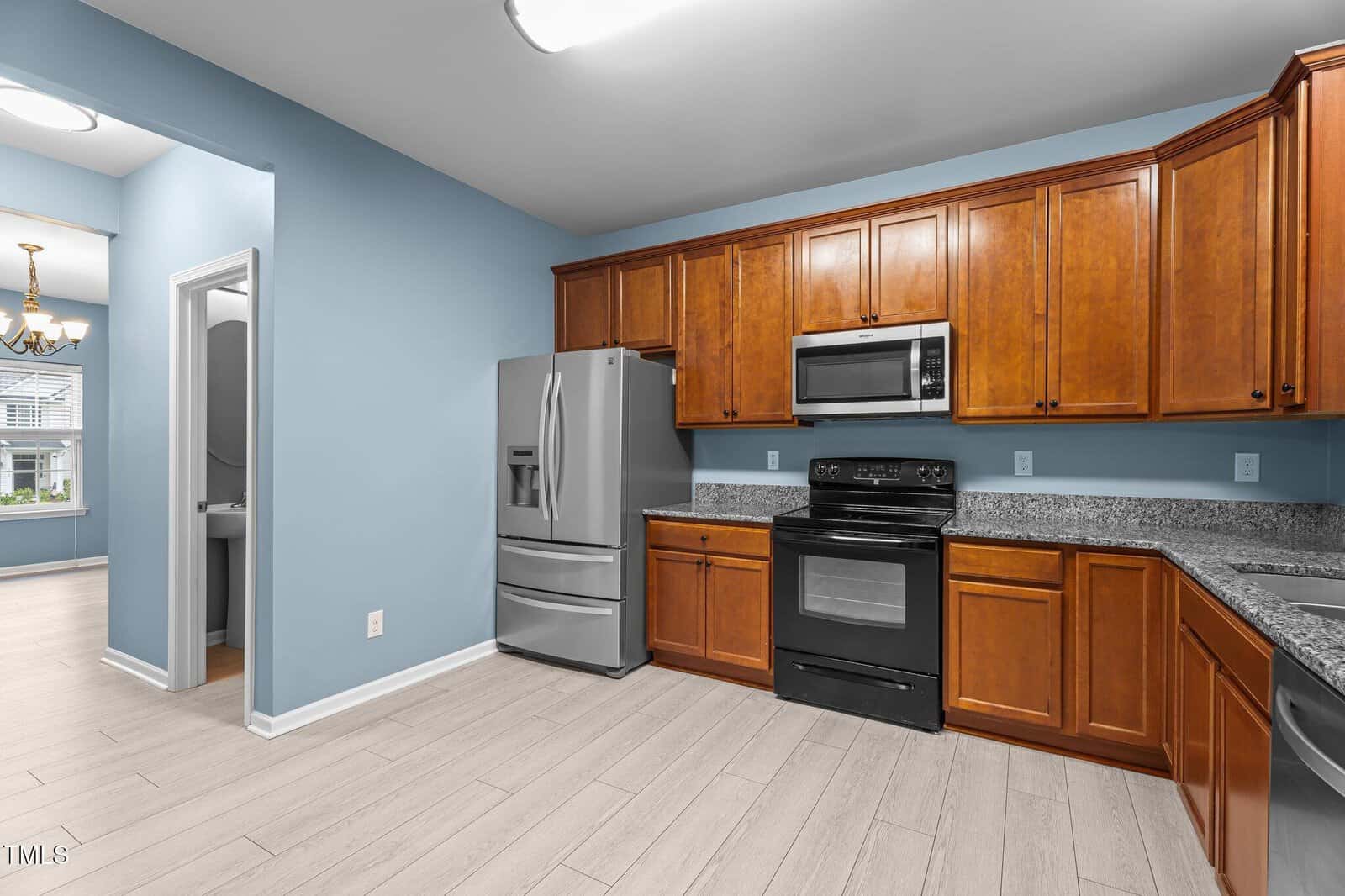 A modern kitchen with light blue walls, light wood flooring, and wooden cabinets. It features a stainless steel refrigerator, a black stove with a microwave above, and a granite countertop. An open doorway leads to a dining area with a chandelier.
