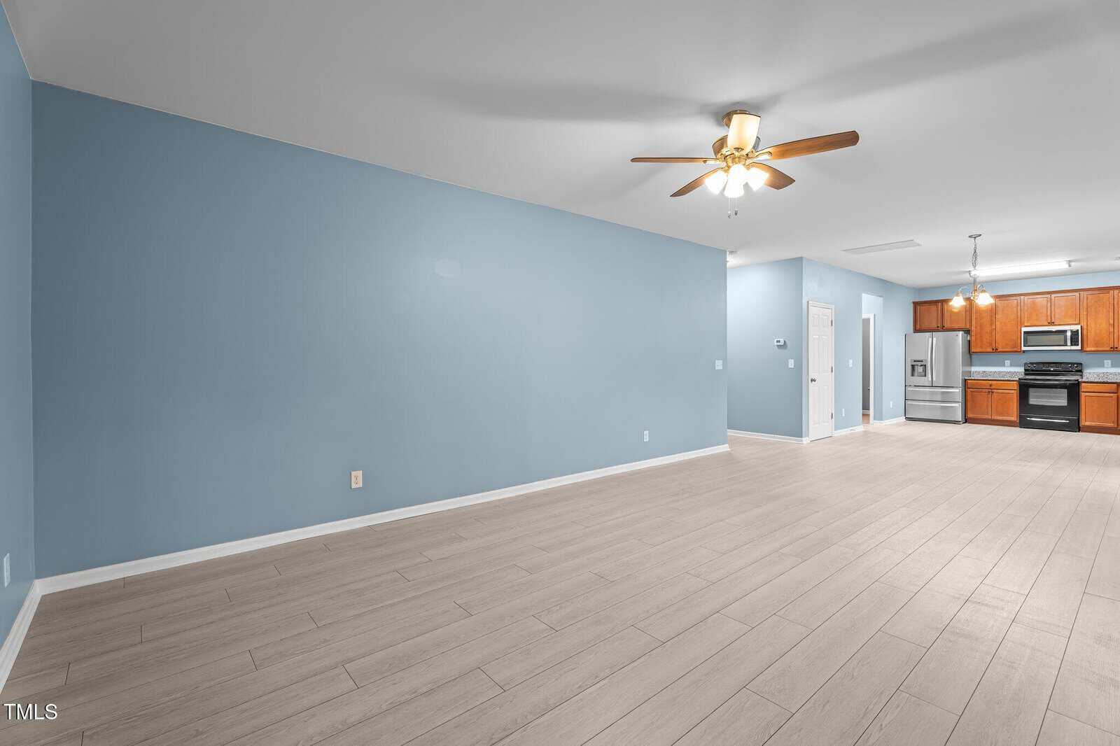 Spacious open-concept living area with light wood flooring and blue walls. Ceiling fan with lights is centered above the room. In the background, a kitchen features wooden cabinets, stainless steel appliances, and two pendant lights.