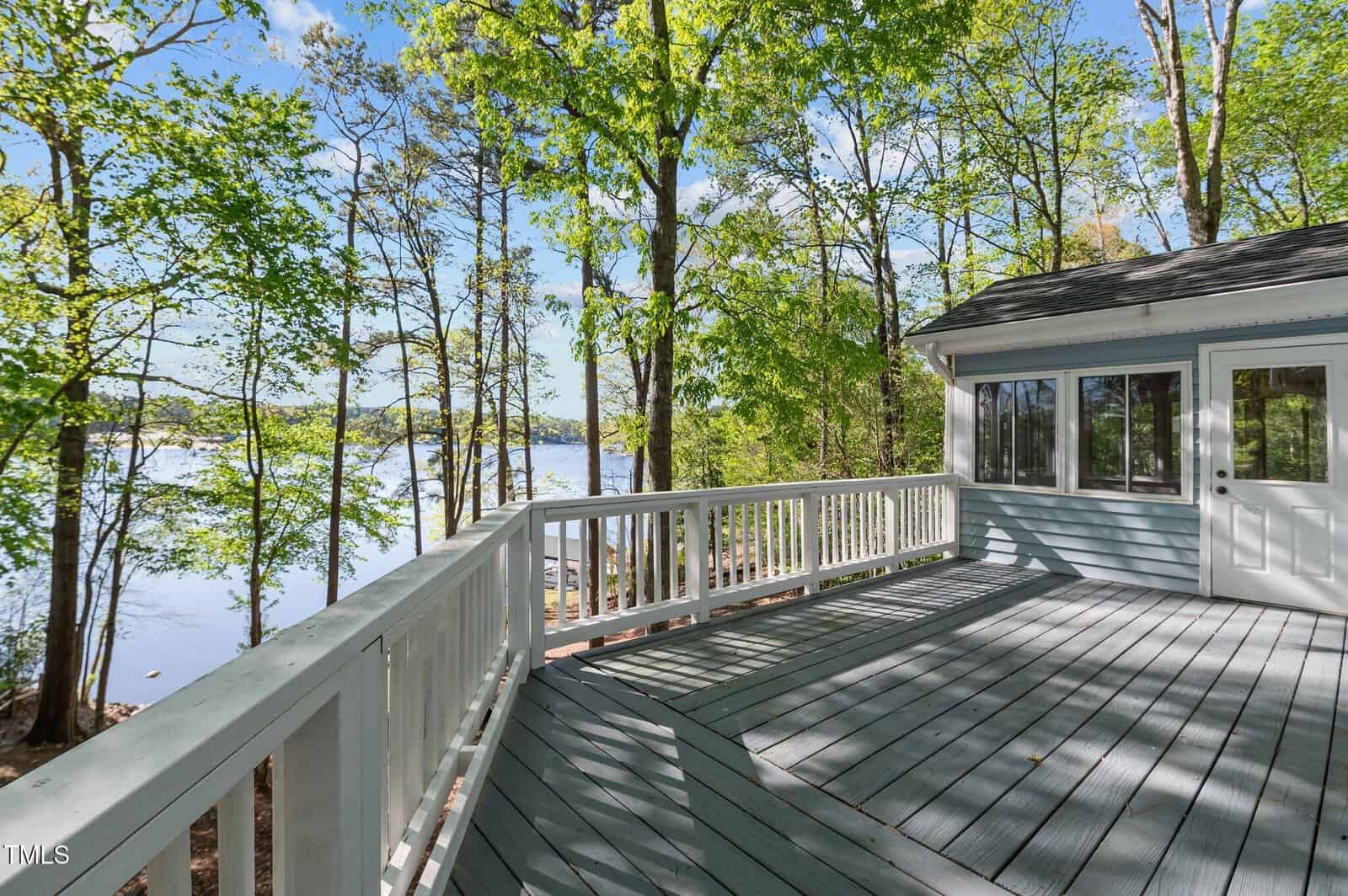 A spacious wooden deck with white railings overlooks a serene lake surrounded by tall trees. The deck is attached to a light blue house with large windows, under a clear, sunny sky.