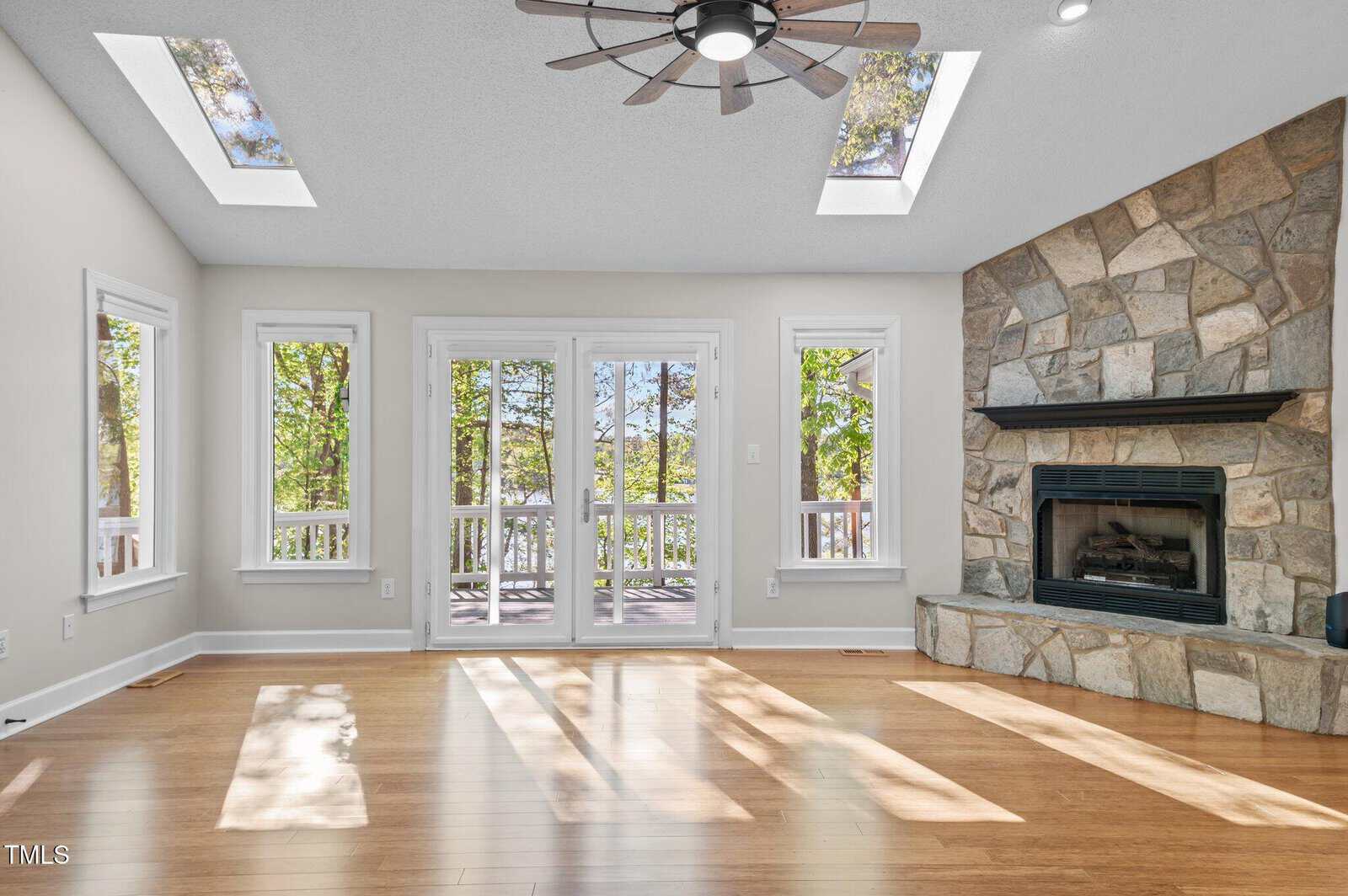 A bright living room with large windows, two skylights, light wood floors, a stone fireplace, and glass doors opening to a deck with trees visible outside. Sunlight streams in, casting shadows on the floor.