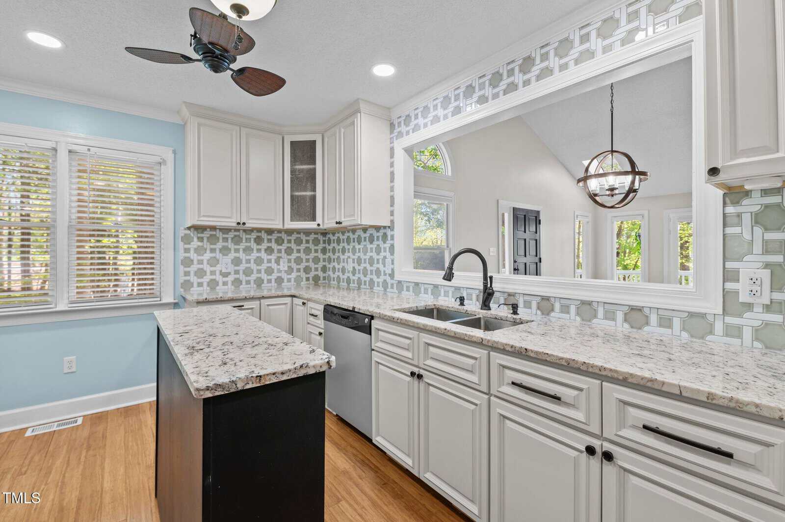 Modern kitchen with white cabinets, patterned tile backsplash, granite countertops, stainless steel appliances, and a large pass-through window. Light blue walls and wood flooring are also visible.