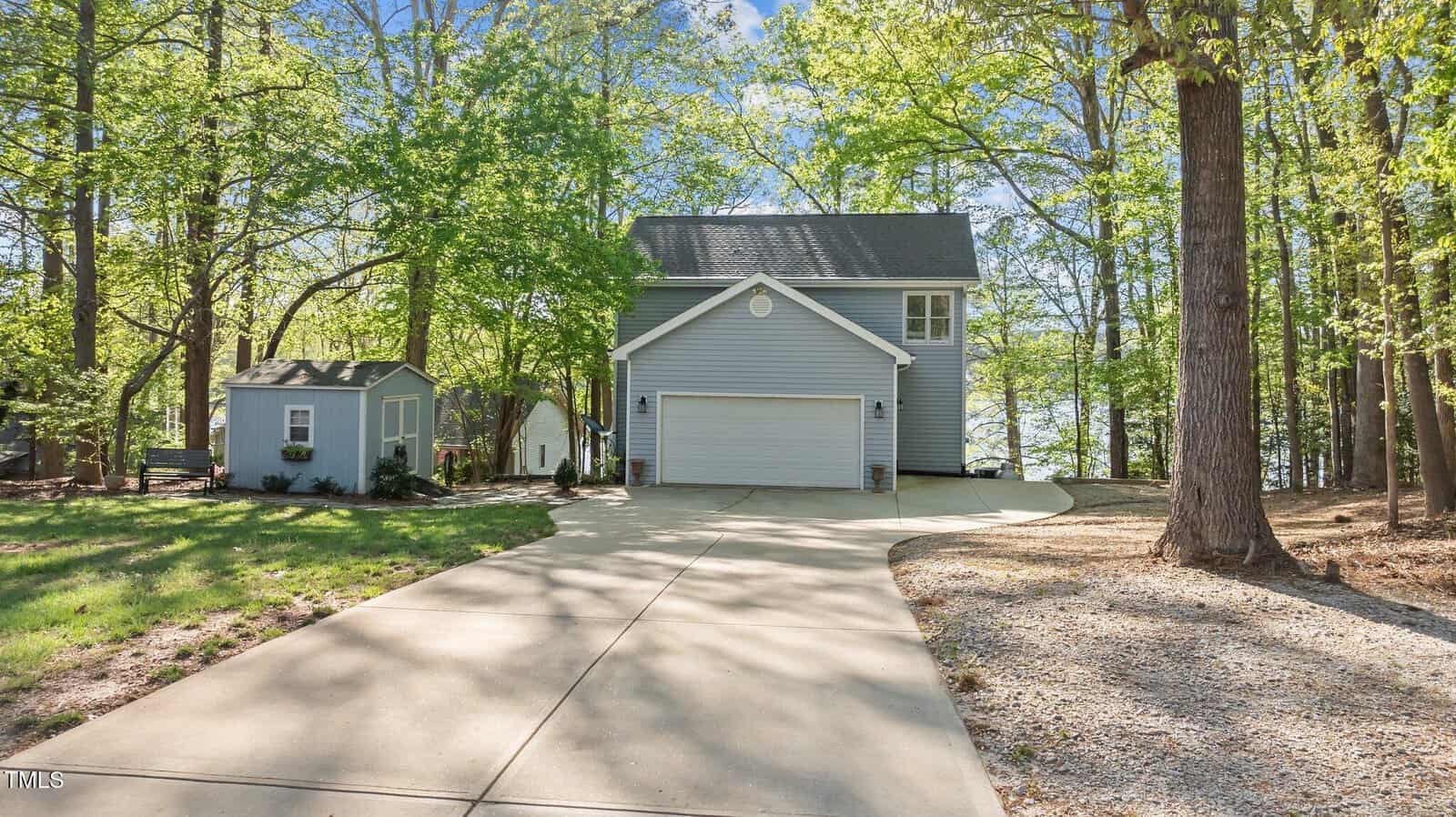A two-story blue house with a white garage sits among tall trees, with a curved driveway leading to it. A small matching shed is to the left, and a lake is visible behind the house under a clear sky.
