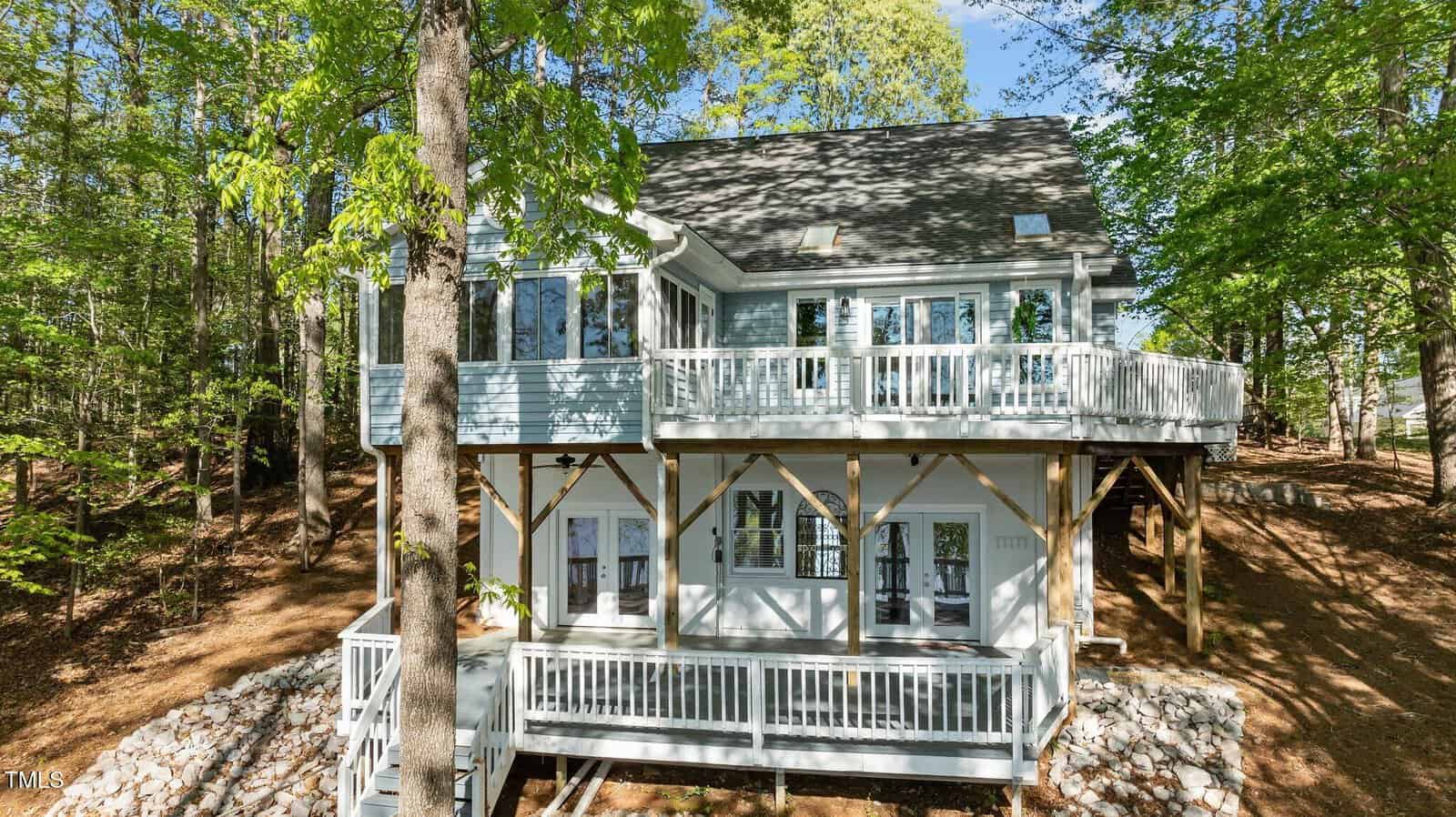 Two-story light blue house with white trim and railings, elevated on stilts, surrounded by trees and a rocky, wooded landscape. The house features two balconies and large windows on both levels.