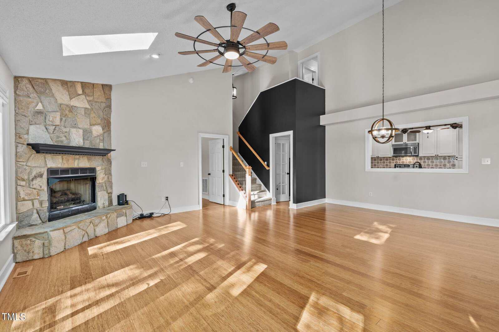 Spacious living room with wood flooring, stone fireplace, large ceiling fan, and high ceiling. Stairs with black accent wall lead upstairs. Open window shows kitchen in the background. Sunlight streams through windows.