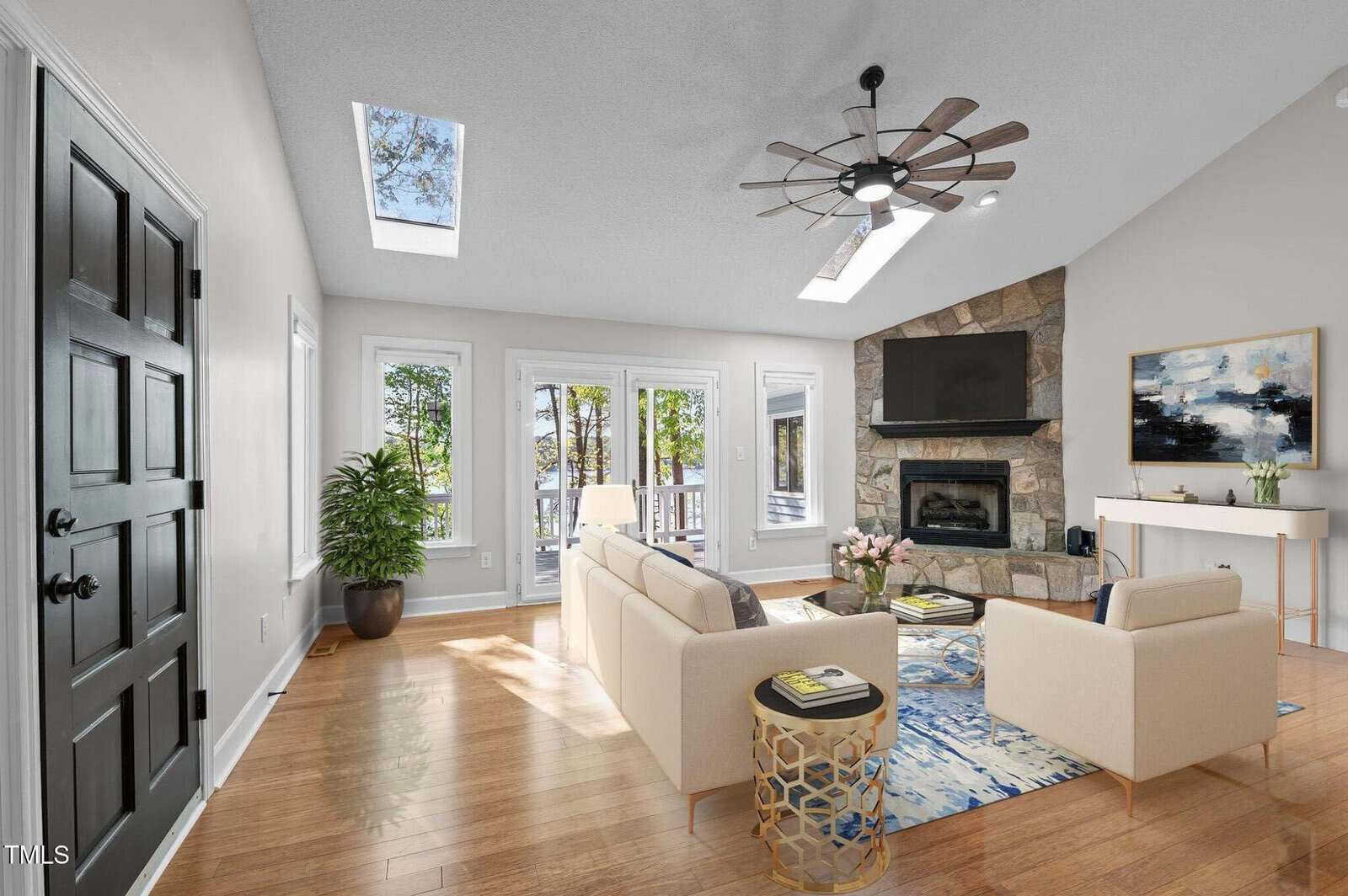 A bright living room with skylights, wood floors, white walls, a beige sofa set, a stone fireplace with a TV above, a ceiling fan, potted plants, and glass doors leading outside. Decor includes a rug and wall art.