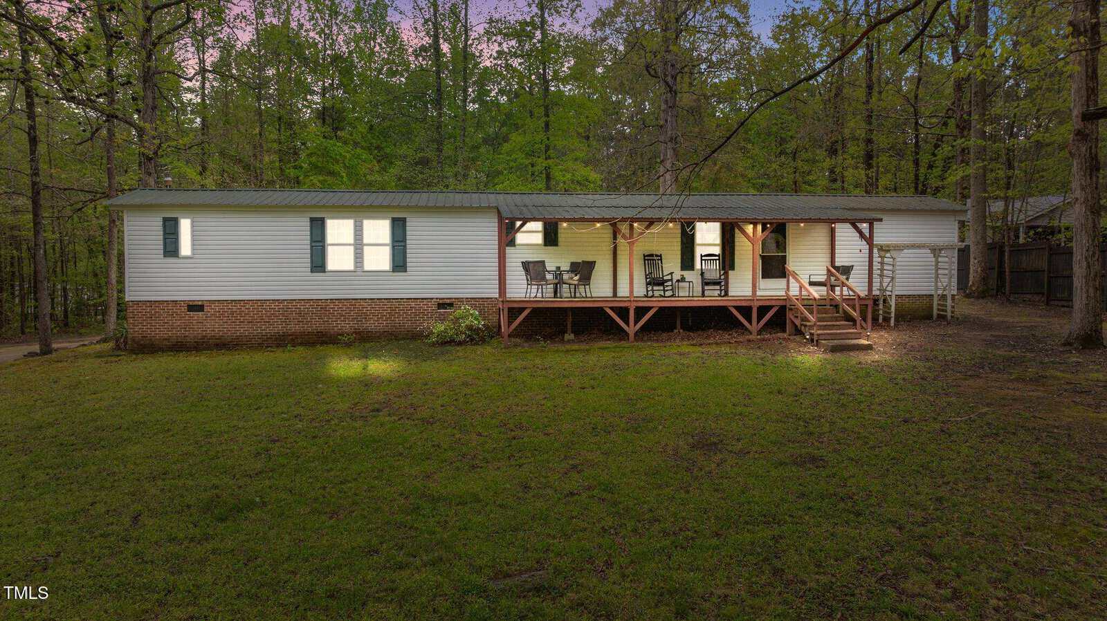 A single-story white manufactured home with green shutters and a brick foundation, featuring a covered wooden porch with outdoor furniture, set in a grassy yard surrounded by tall trees.
