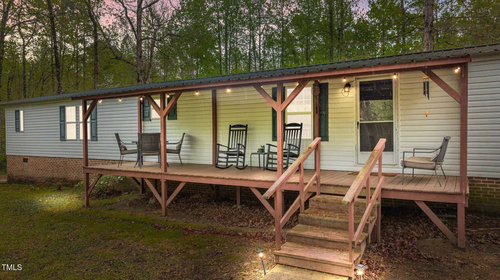 A single-story white manufactured home with a covered wooden porch, three sets of chairs and tables, a string of lights, steps leading up, and surrounded by trees in the evening.
