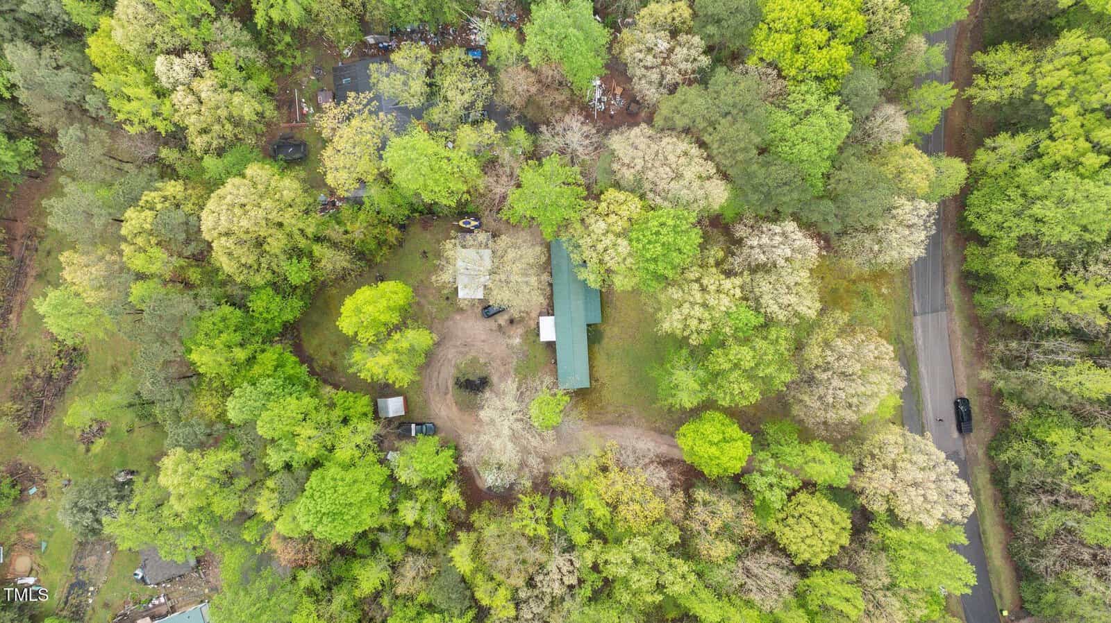 Aerial view of a rural property surrounded by dense green trees, featuring a long, green-roofed building, a driveway, several smaller structures, and a paved road with a vehicle passing by on the right side.