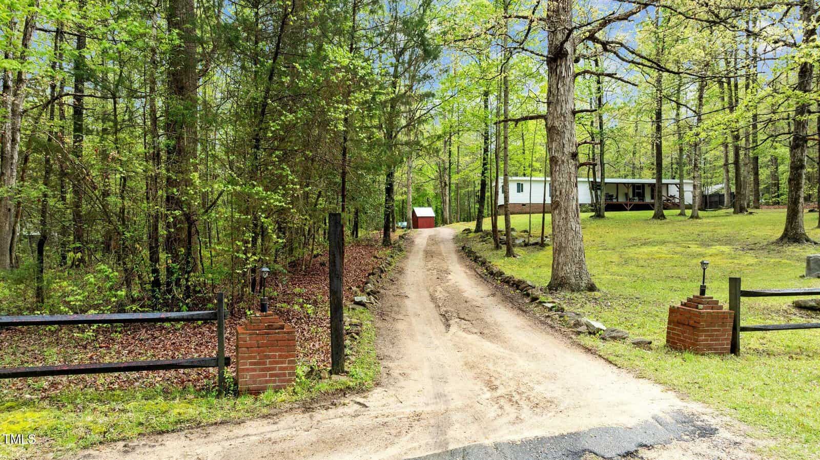 A dirt driveway leads through a wooded area with a broken brick fence at the entrance, ending at a white single-story house with a porch and a red shed in the background. Trees and greenery surround the property.