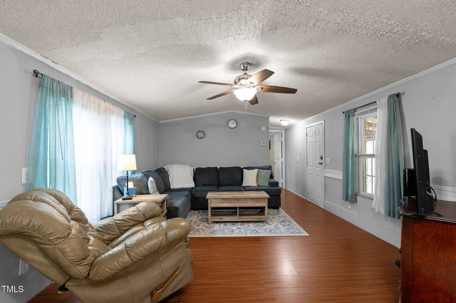 A living room with wood flooring, a brown recliner, a sectional sofa, a coffee table, a TV on a stand, two windows with teal curtains, and a ceiling fan with a light fixture.