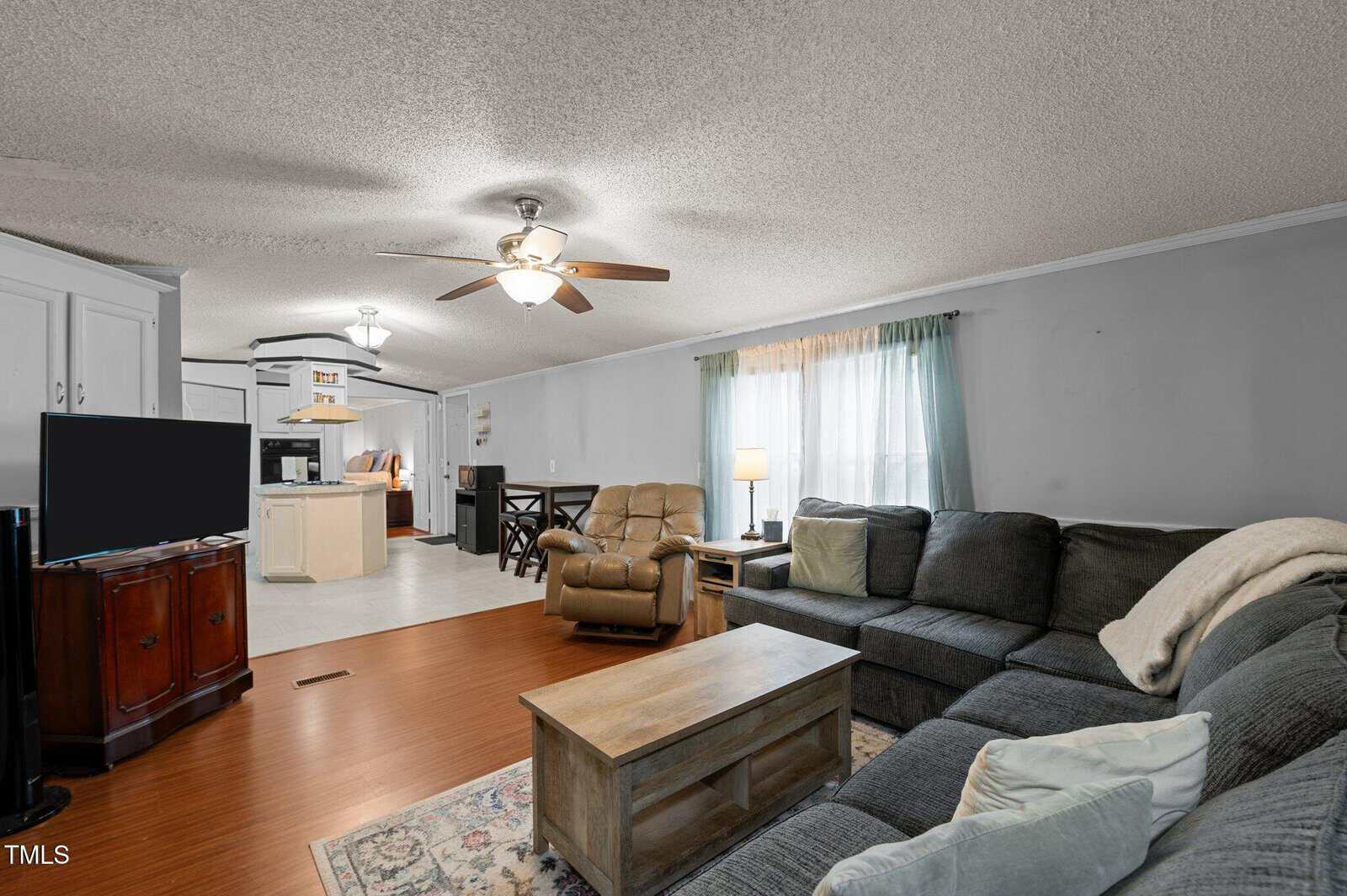 A living room with a gray sectional sofa, coffee table, recliner, TV on a stand, and a rug. The space connects to a kitchen and dining area. Light blue curtains cover the windows. Ceiling fan and lamp provide lighting.