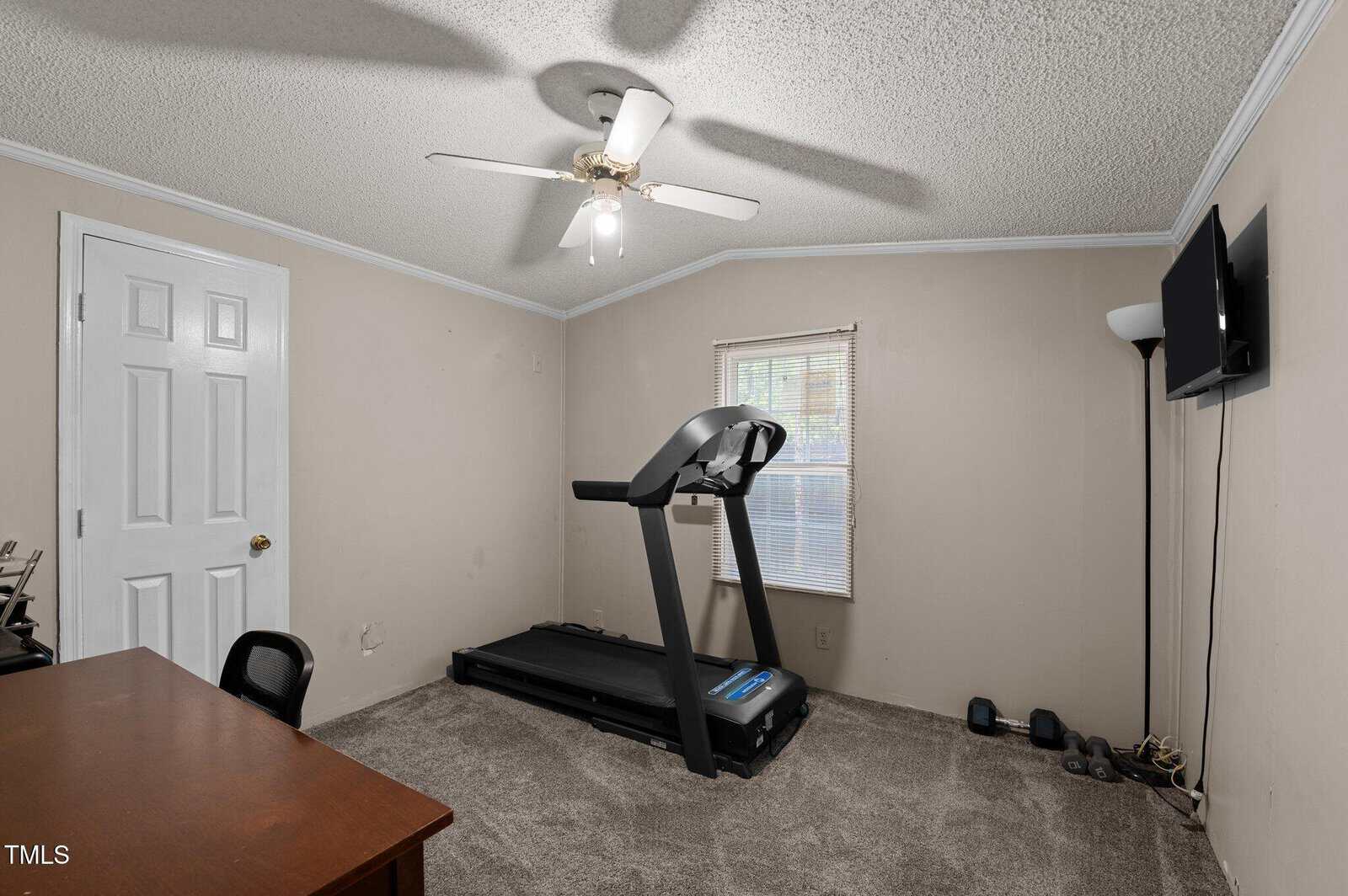 A small room with beige walls and carpet, containing a treadmill, a desk with a chair, a mounted TV, a floor lamp, and some dumbbells and shoes near the window. Ceiling fan with lights is on the ceiling.