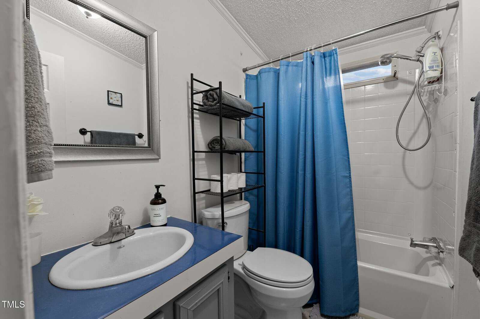 A small bathroom with a blue countertop sink, mirror, metal shelving unit over the toilet, white tiles in the shower, blue shower curtain, towels, and a wall-mounted showerhead.