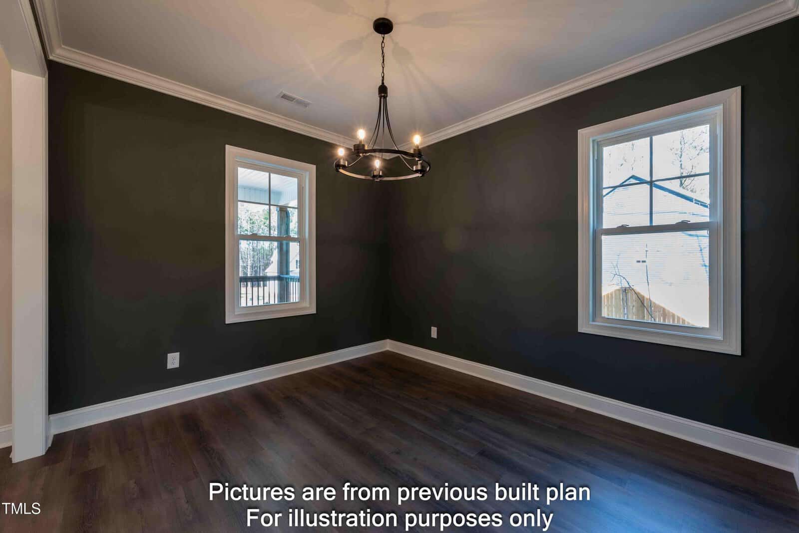 An empty room with dark green walls, two windows, hardwood floors, crown molding, and a modern black chandelier. Sunlight enters through the windows. Text at the bottom says, “Pictures are from previous built plan for illustration purposes only.”.