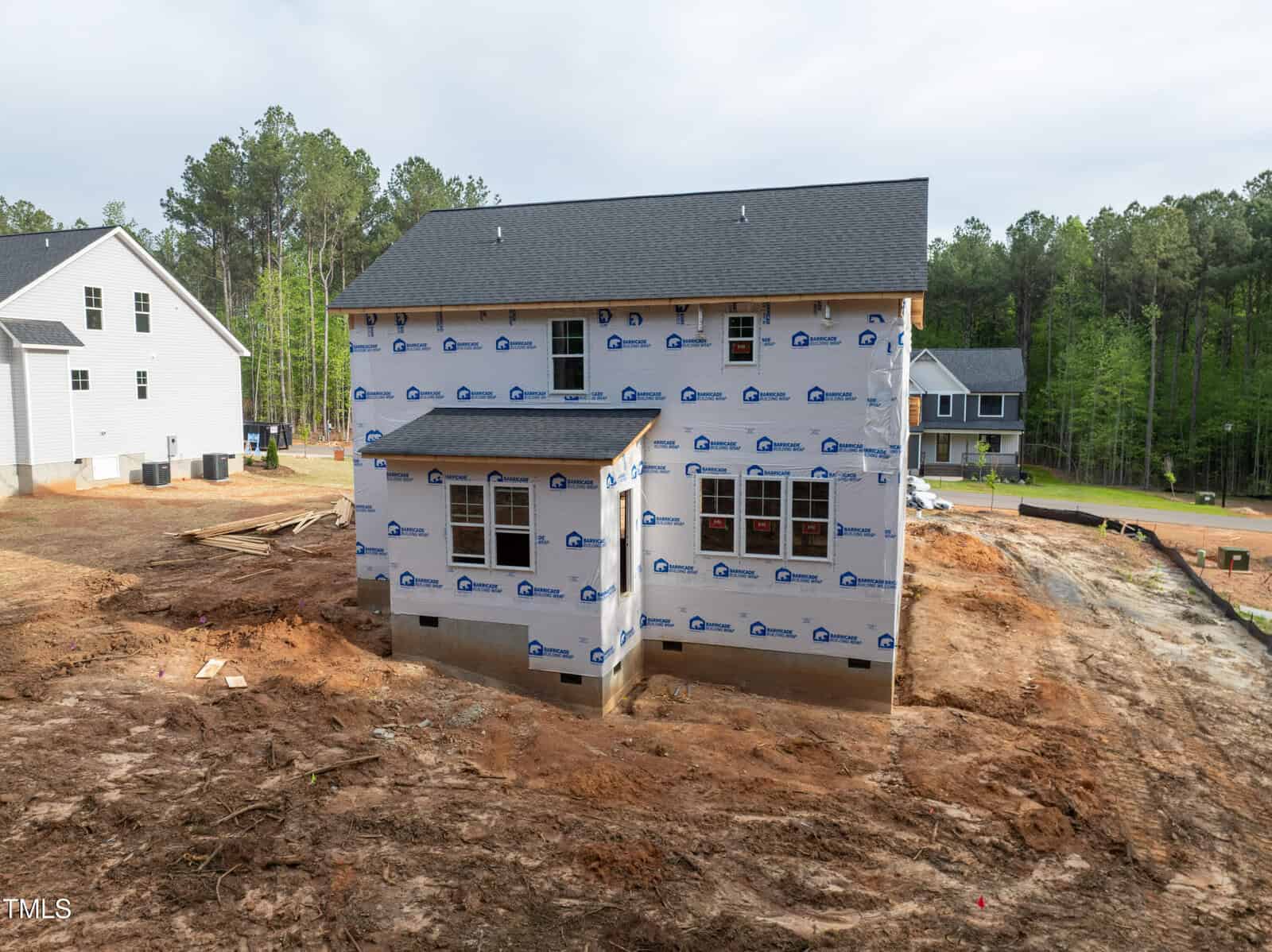 A two-story house under construction with exterior walls covered in weather-resistant wrap, surrounded by bare earth and some construction materials, with other completed houses and trees in the background.