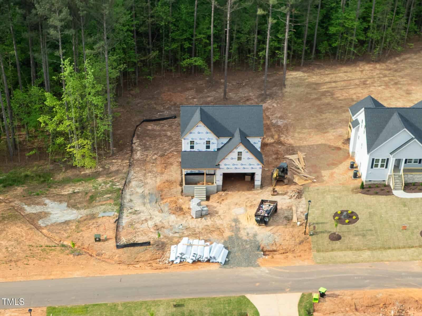 Aerial view of a two-story house under construction with exposed framing and building materials scattered in the yard, next to a completed house, bordered by trees and a cleared dirt lot.