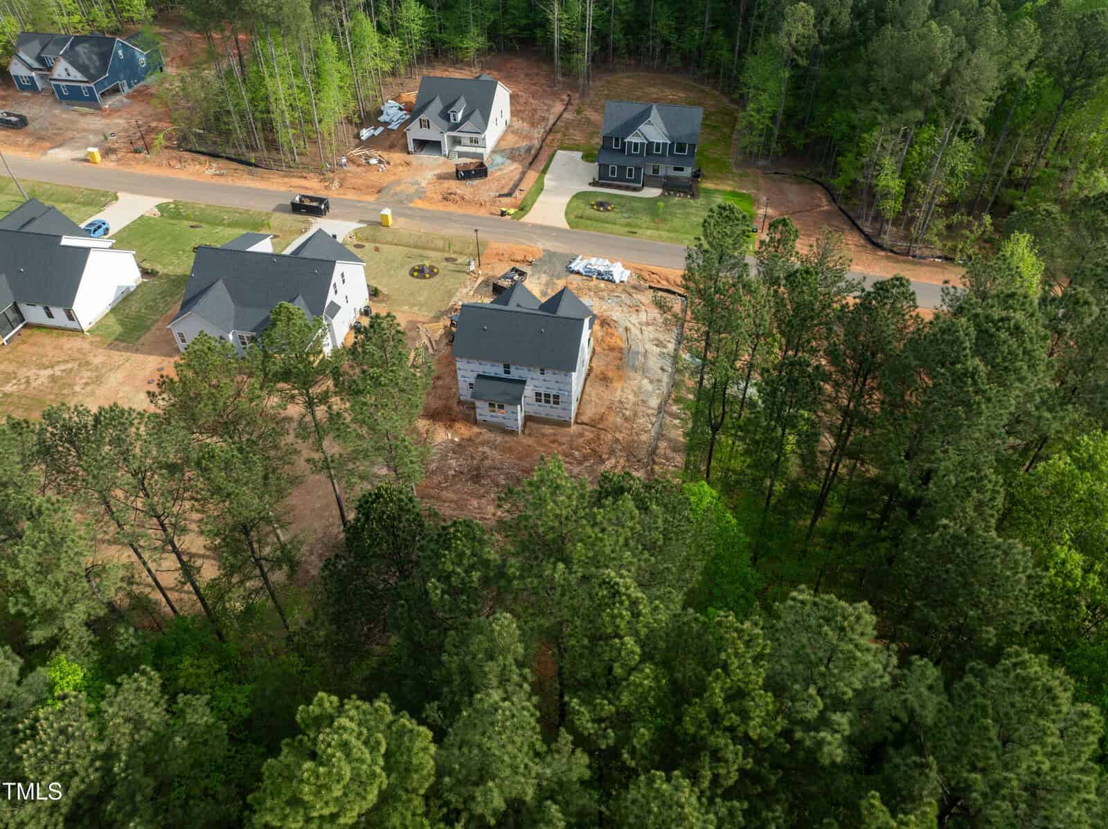 Aerial view of a residential neighborhood with several houses surrounded by trees. One house is under construction with cleared land around it, while other completed homes are nearby along a paved road.