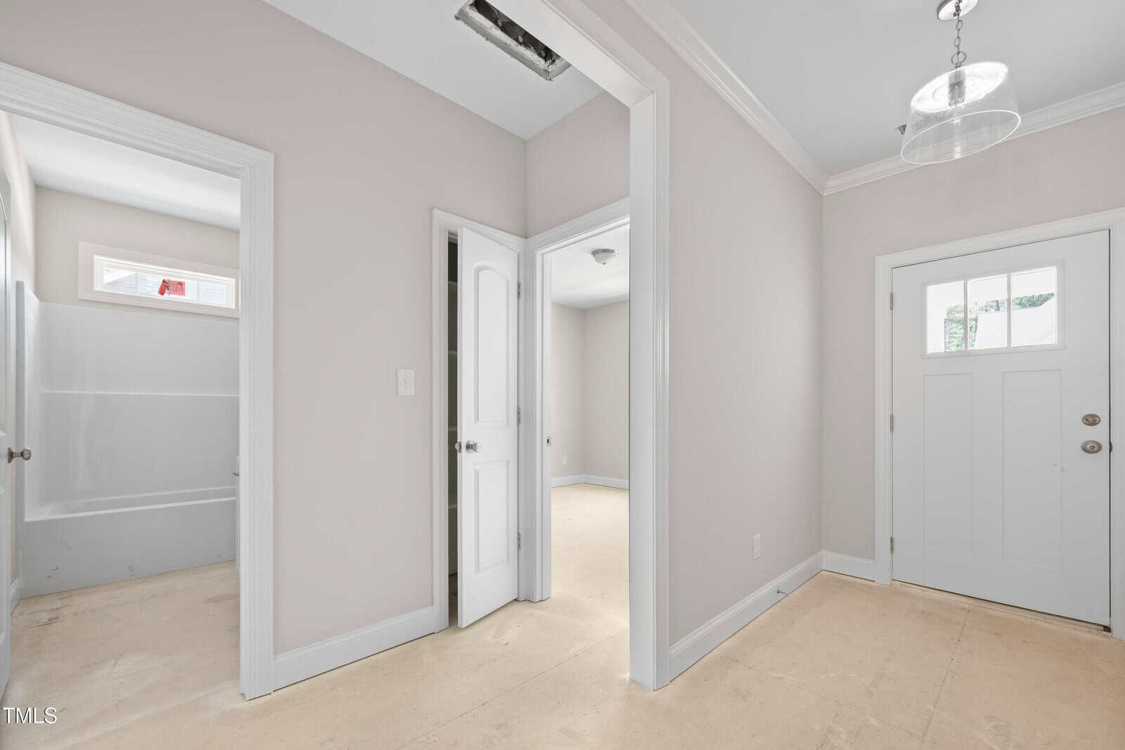 A hallway with beige walls and unfinished floors, showing a white door with glass panels, two open doorways leading to other rooms, and a bathroom with a bathtub visible. Ceiling lights are installed.