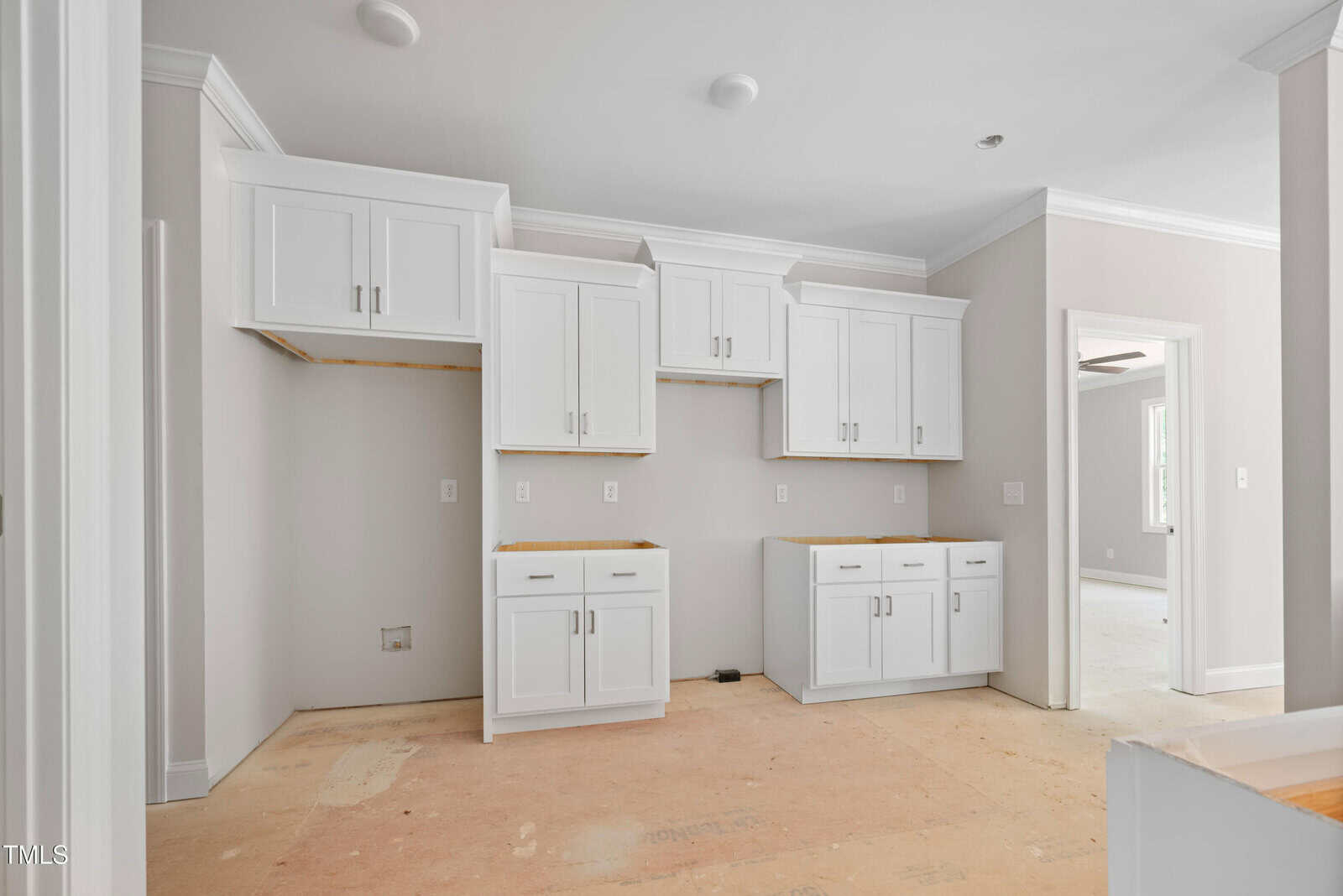 A kitchen under construction with white cabinets installed, but missing appliances, countertops, and hardware. The floor is unfinished, and walls are painted light beige. An adjacent room with a ceiling fan is visible in the background.