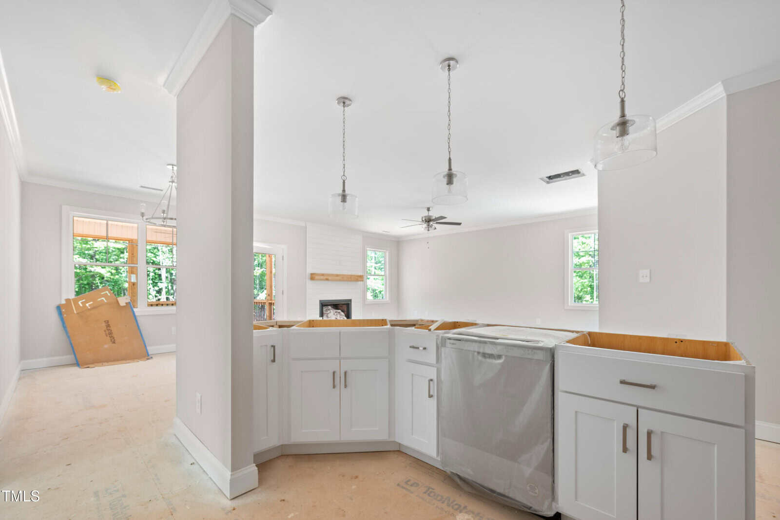 A modern kitchen under construction with white cabinets, a dishwasher installed, pendant lights, unfinished flooring, and an open view to a living area with large windows and a fireplace.
