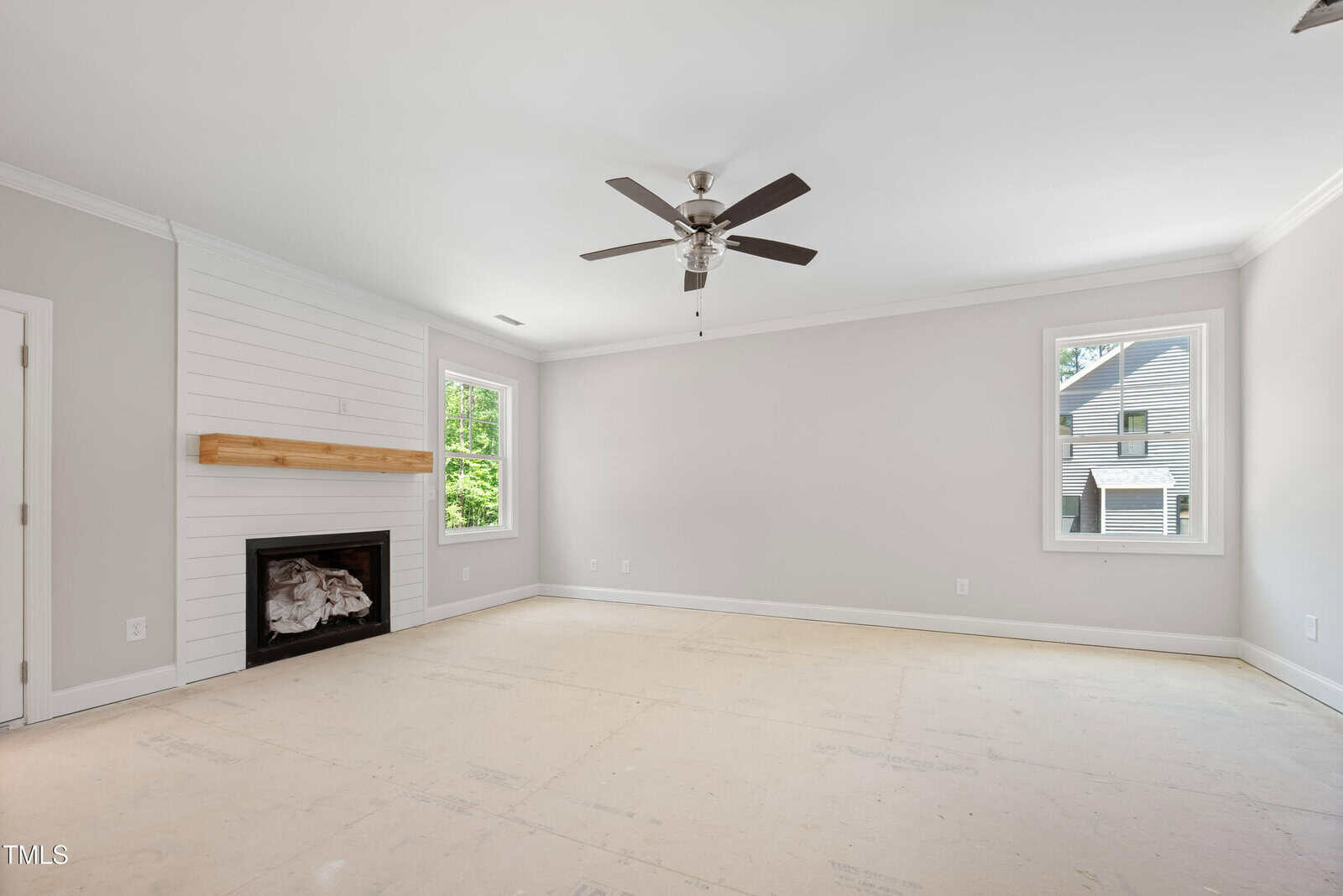 Empty, unfurnished room with light walls, two windows, a ceiling fan with lights, and a fireplace with a wooden mantel. The floor appears unfinished. Natural light comes in from two windows on adjacent walls.