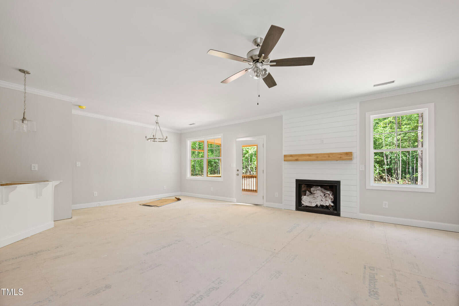 Empty living room with white walls, a ceiling fan, fireplace with wooden mantel, large windows, and a door leading outside. A chandelier is over the dining area, and the floor is unfinished. Trees are visible through the windows.