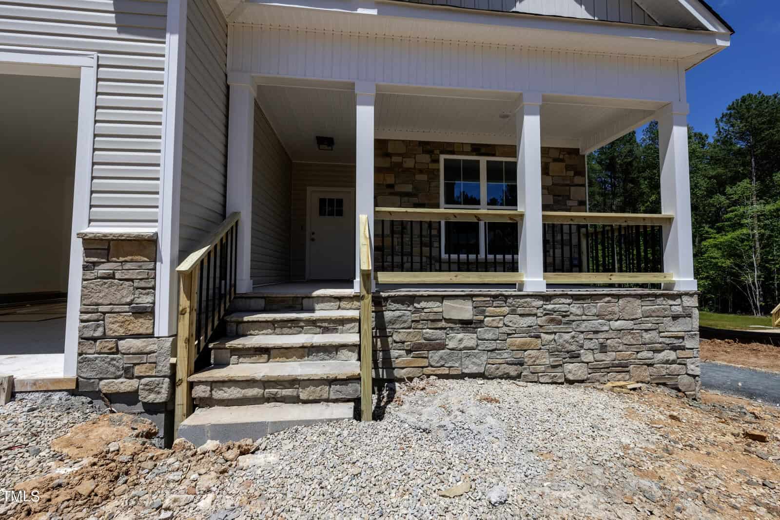 A newly constructed house with a front porch featuring stone and wood accents, a set of stairs leading to the entryway, and unfinished landscaping with exposed dirt and gravel in the foreground.