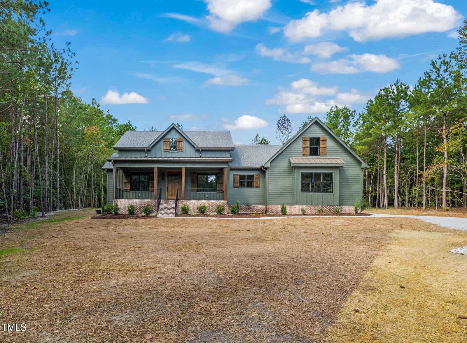 A green single-story house with a covered front porch, dormer windows, and a gable roof, surrounded by pine trees and a grassy yard under a partly cloudy sky.