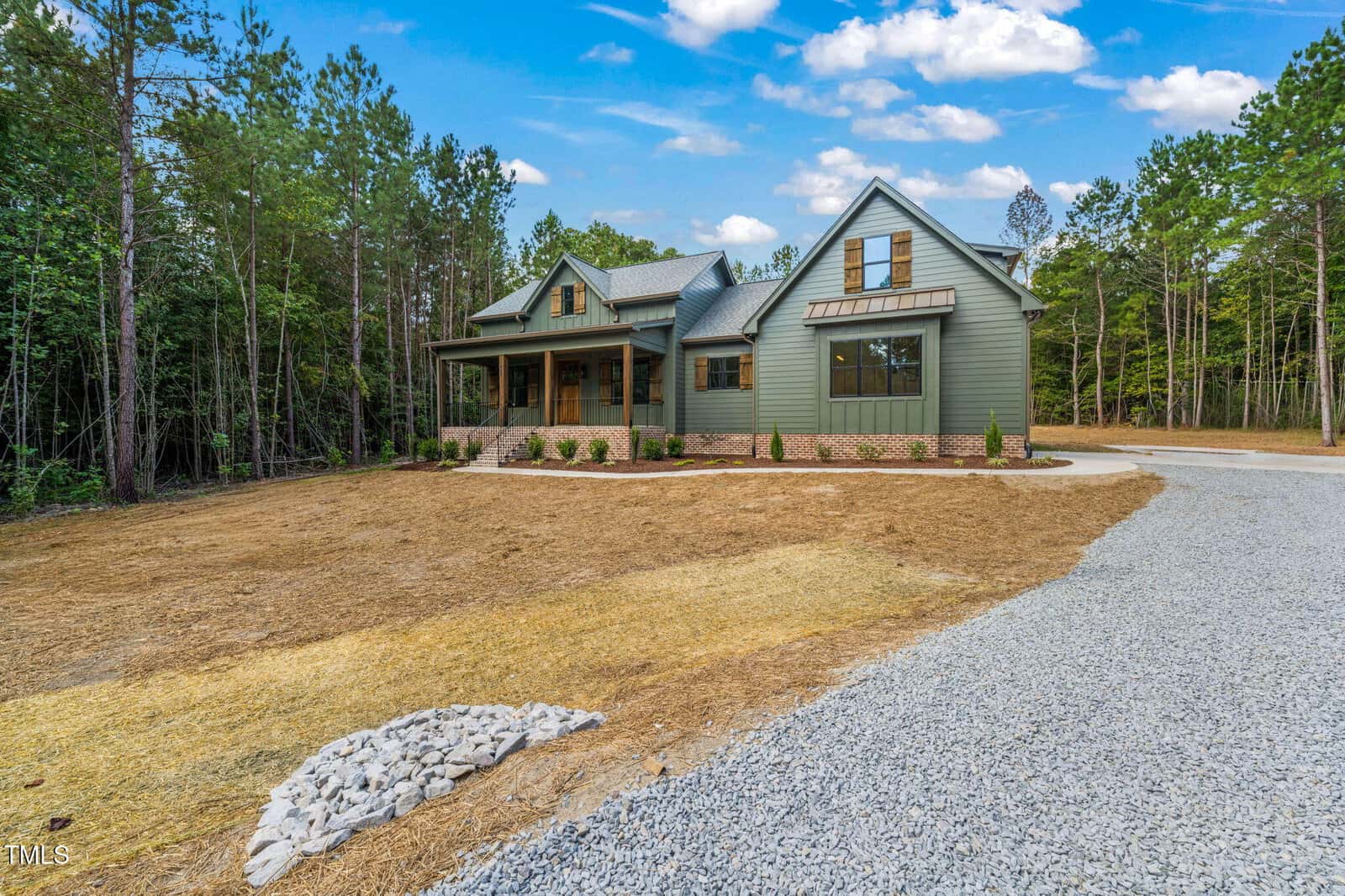 A green, two-story house with a front porch sits next to a gravel driveway surrounded by trees and a partially grassed yard under a blue sky with scattered clouds.