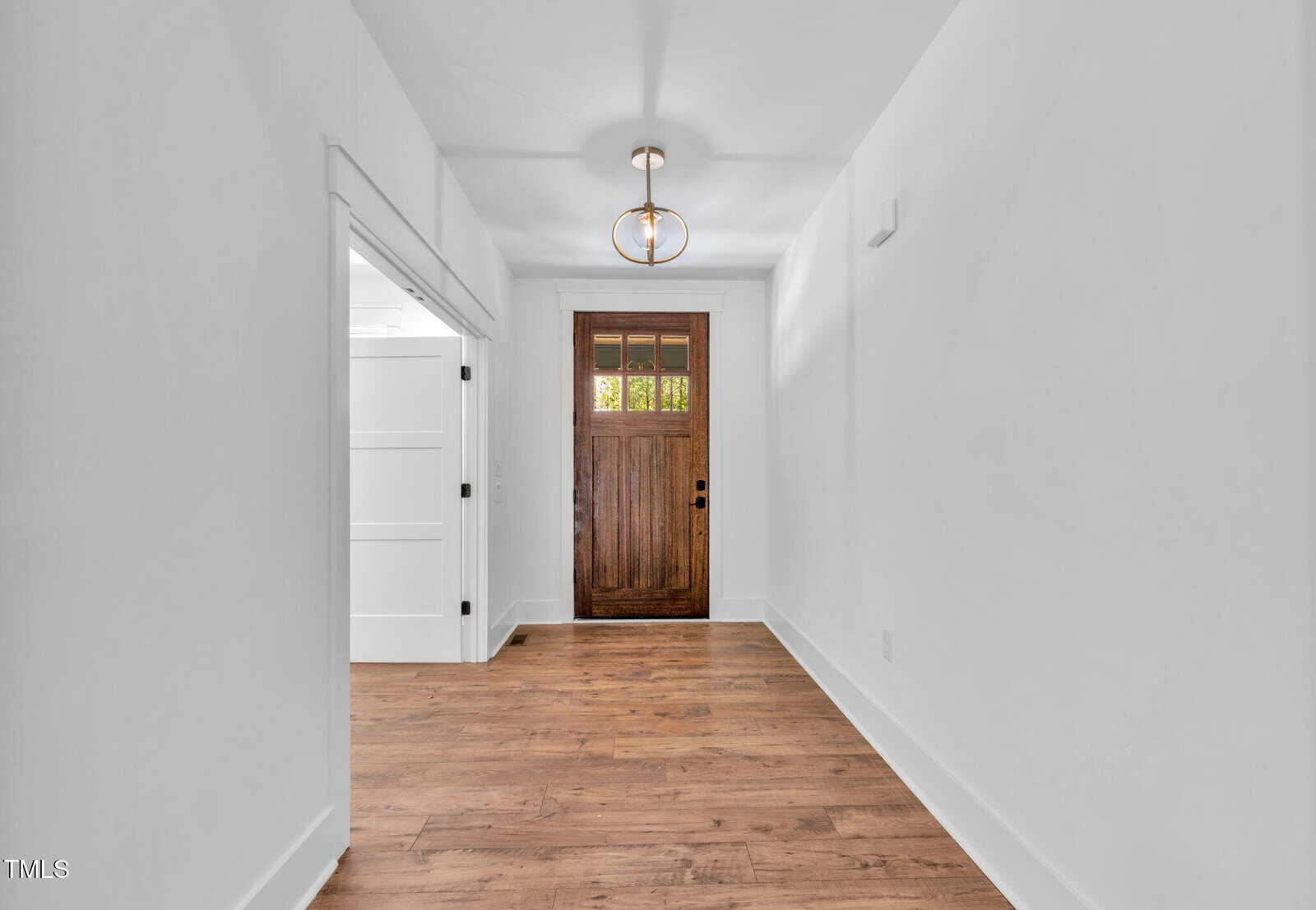 A hallway with white walls and ceiling, wooden flooring, a wooden front door with glass panes at the top, and a modern ceiling light fixture. An open white double door is visible on the left side.