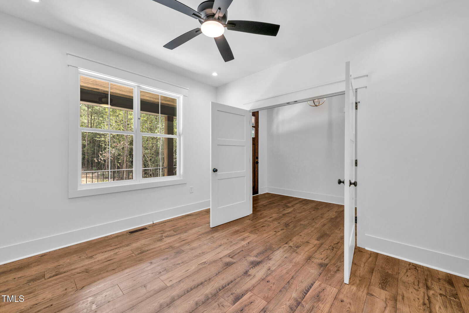 A bright, empty room with white walls, double white doors, a window overlooking trees, a ceiling fan with lights, and wood flooring. Natural light comes in through the window.