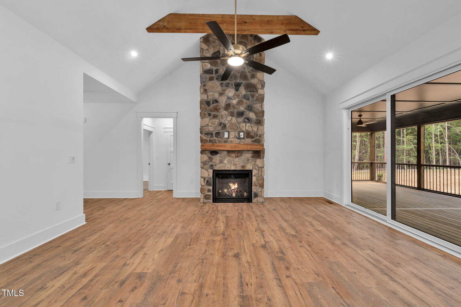 Spacious living room with vaulted ceiling, a stone fireplace with a wooden mantle, wood flooring, white walls, a ceiling fan, and large sliding glass doors leading to a screened-in porch with a wooded view.
