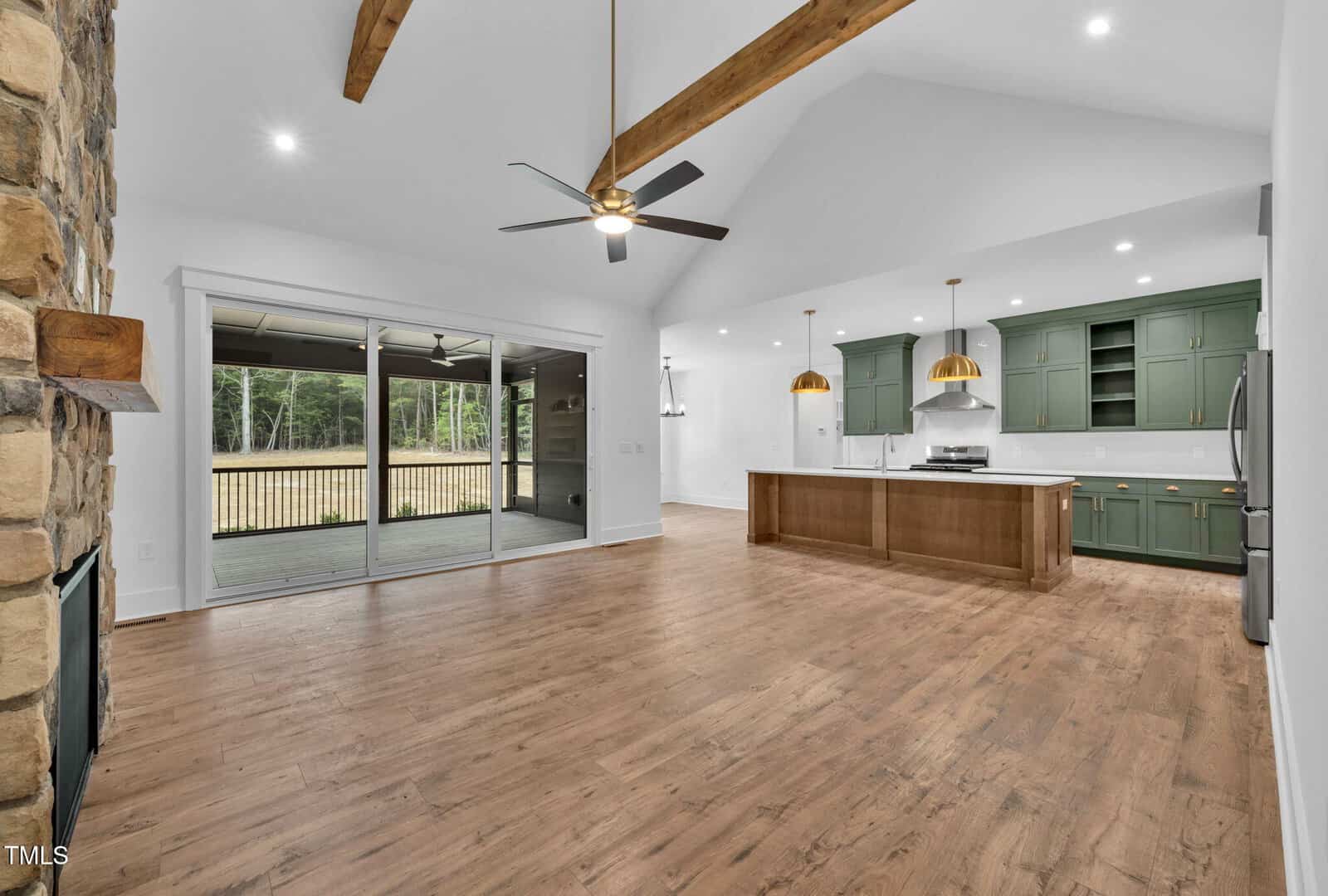 Open-concept living area with wood floors, exposed wooden ceiling beams, a stone fireplace, green kitchen cabinets, an island with pendant lights, stainless steel appliances, and sliding glass doors to a covered patio.