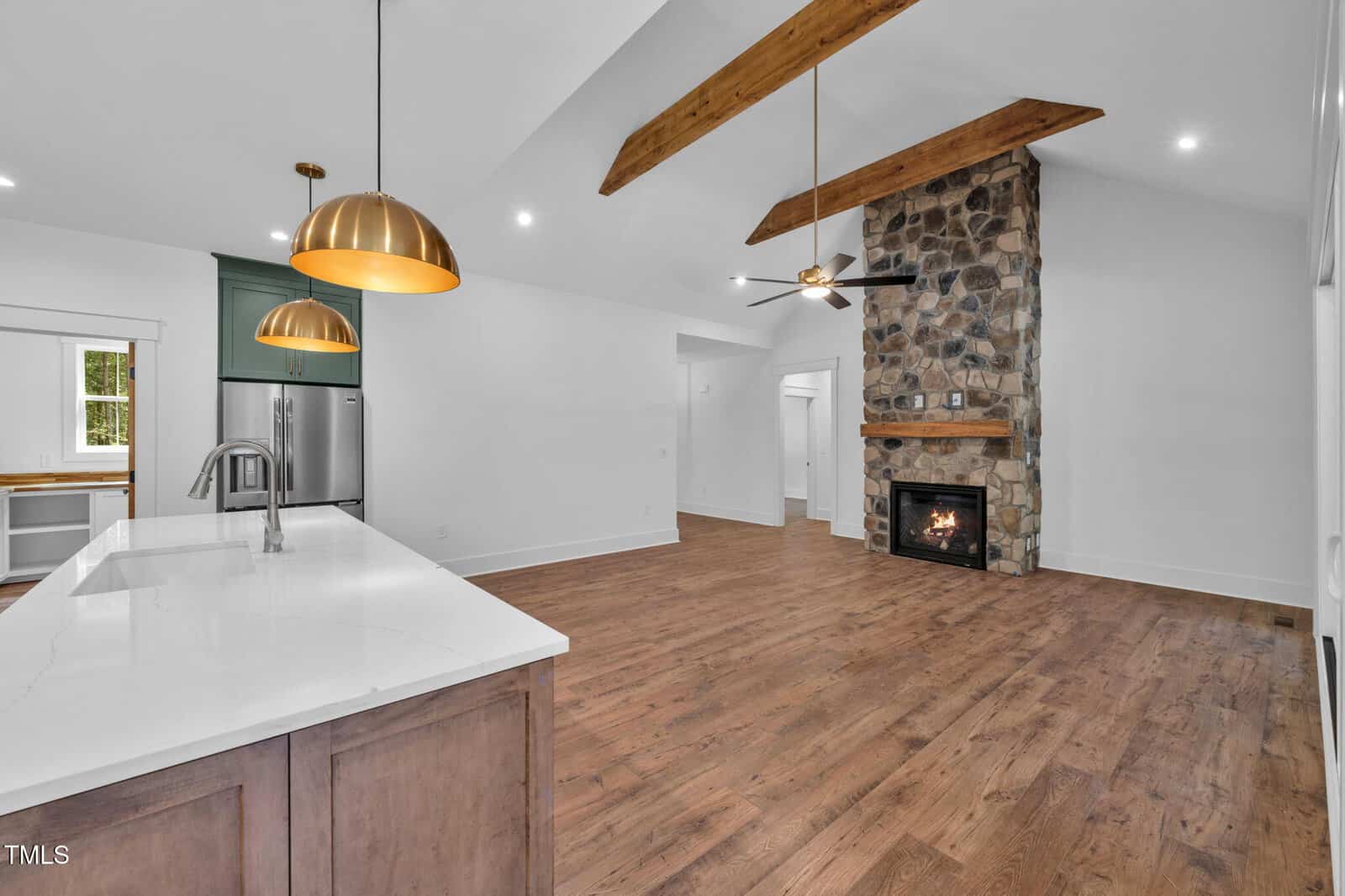Open kitchen and living area with wood floors, white walls, exposed wood beams, a stone fireplace, and modern pendant lights over a large island with a sink. Stainless steel appliances are visible in the background.