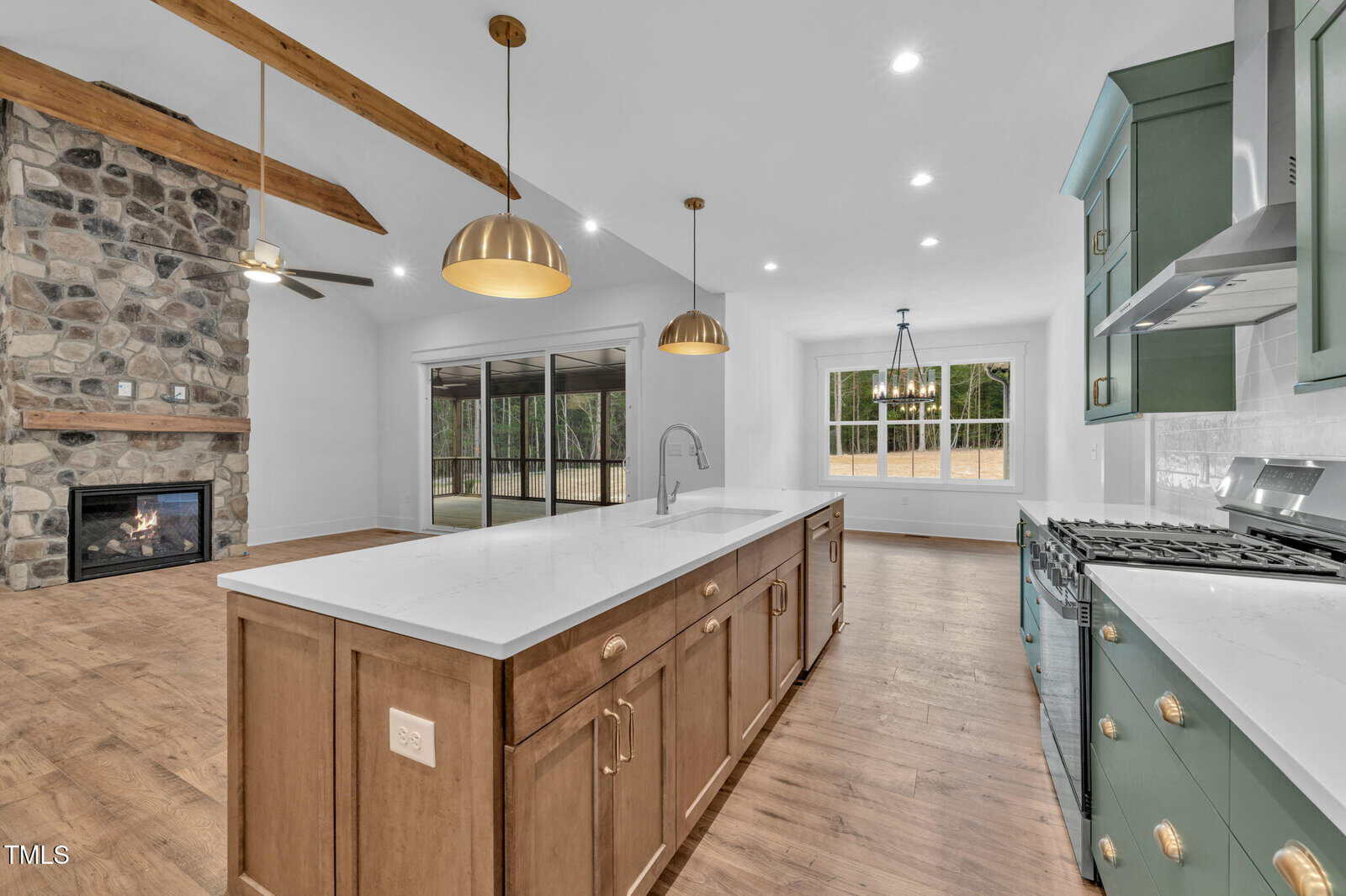 Modern kitchen with a large island, green cabinets, stainless steel appliances, and gold pendant lights. The open layout shows a stone fireplace, ceiling beams, and sliding glass doors leading to a patio.