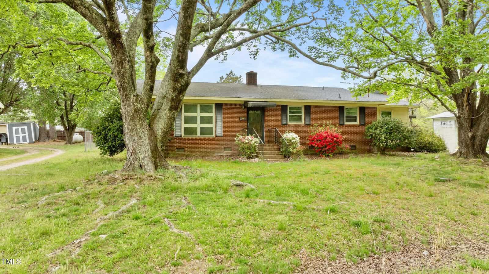 Charming single-story red brick house with a black roof, central door, and multiple windows situated at 428 Mitchell Avenue in Franklinton. Large trees and bushes surround the yard, while vibrant red and white flowers bloom in front of the house. A driveway is visible on the left.