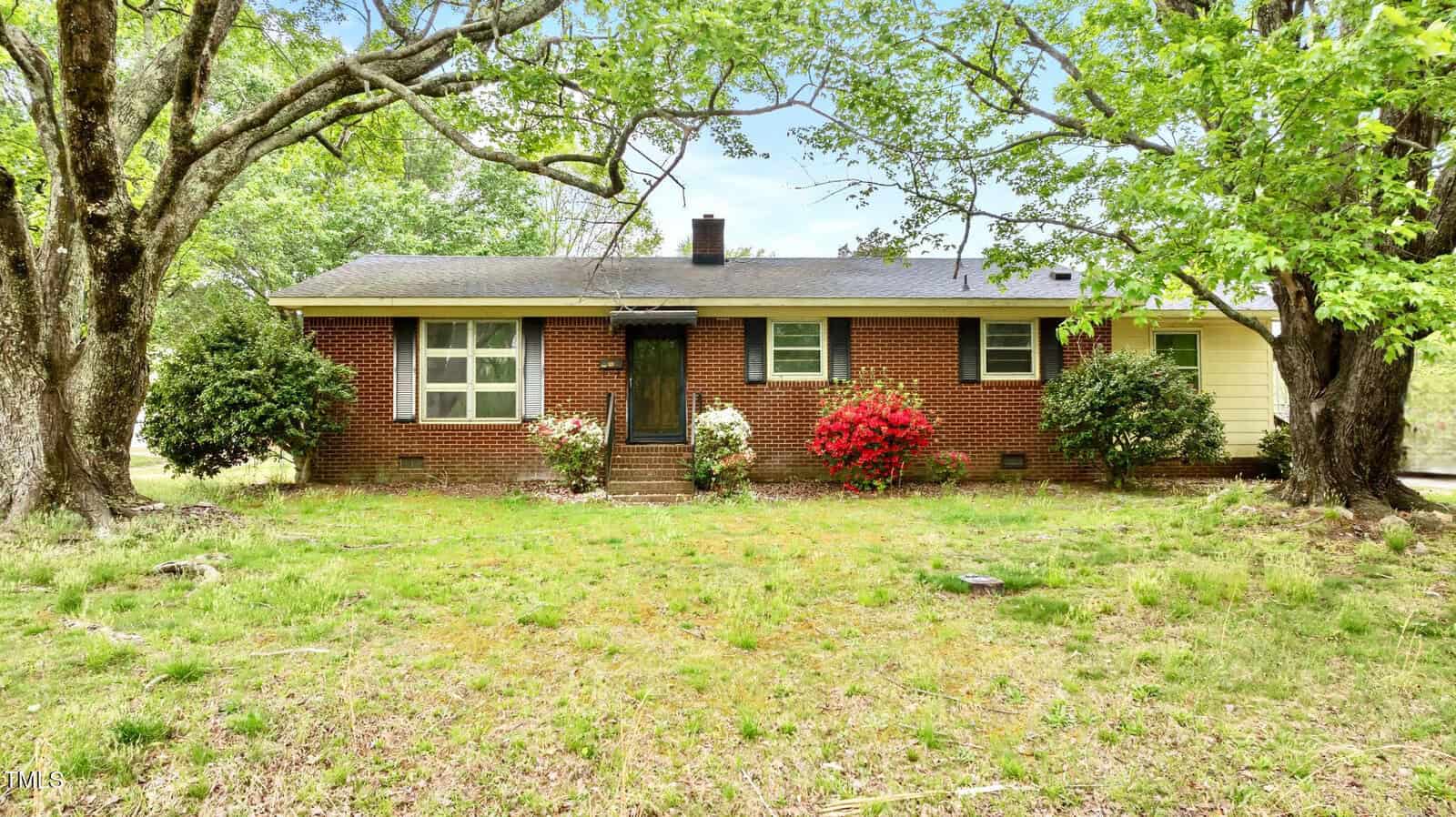 A one-story brick house with a dark roof is surrounded by trees and bushes. There are three windows, a front door, and flowering shrubs in front of the house. The yard has patchy grass and a natural, untended look.