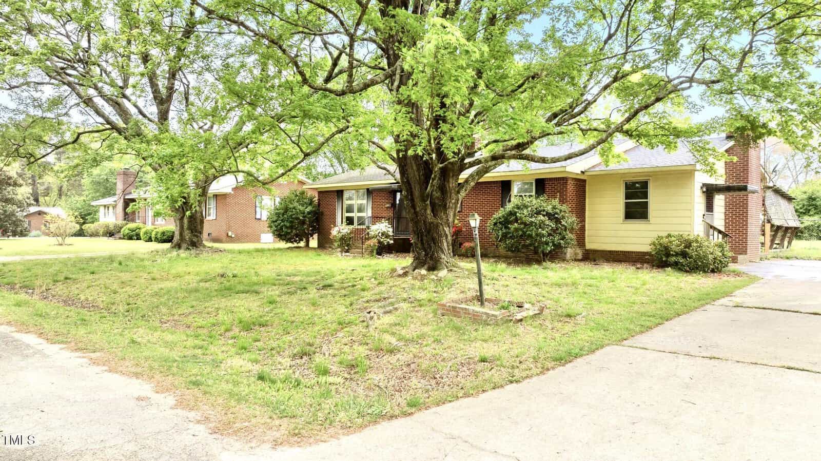 One-story brick house with yellow siding on part of the exterior, a large tree in the front yard, driveway on the right, and a grassy lawn with some shrubs and bushes. Another brick house is visible in the background.