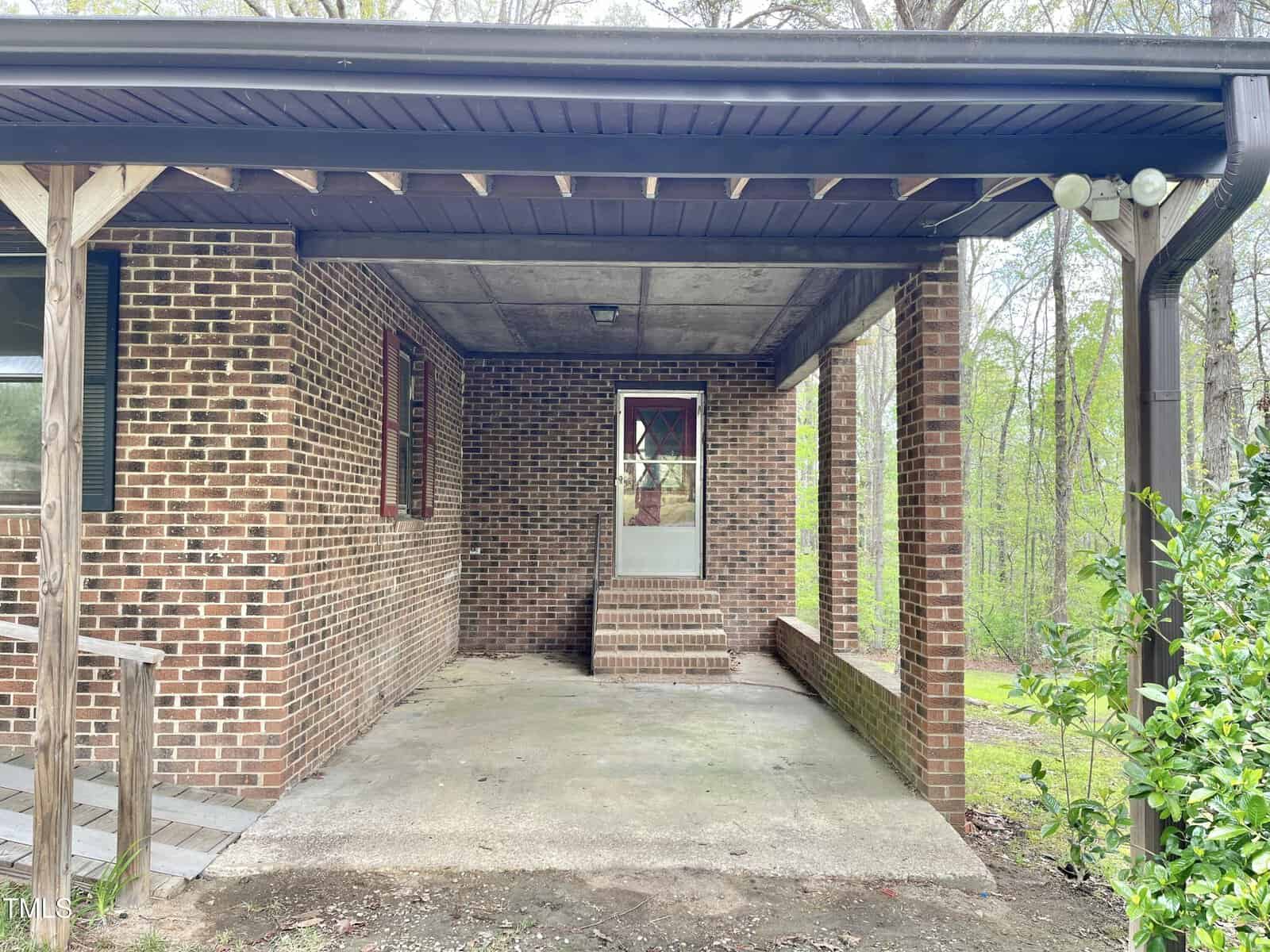 Covered brick porch with concrete floor, steps leading to a door with a window, and red-trimmed windows. Surrounded by trees and greenery in the background. Wooden railing is visible on the left side.