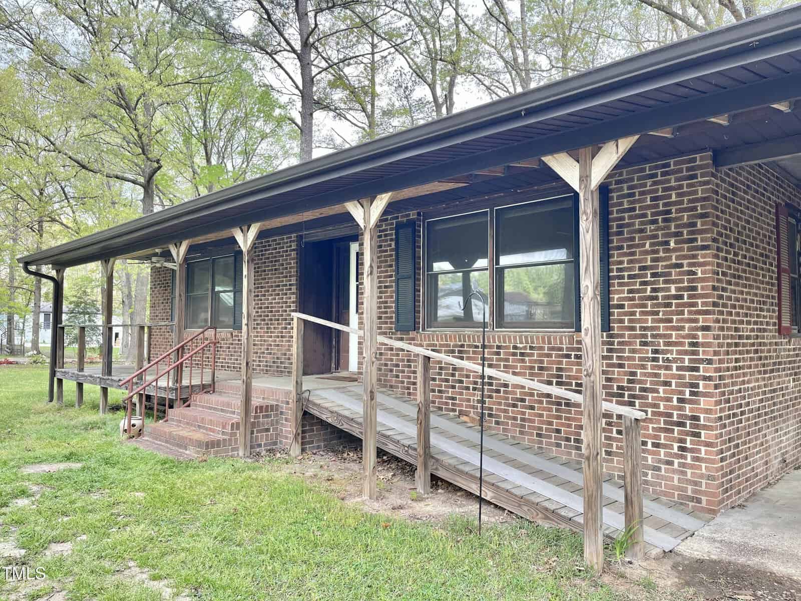 Single-story brick house with a covered front porch, black-framed windows, wooden supports, a wheelchair-accessible ramp, and steps leading to the entrance. Green grass and tall trees surround the house.