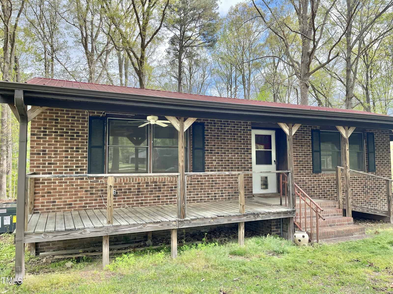 A single-story brick house with a covered front porch, wooden railing, black shutters, and a red metal roof. The porch has a ceiling fan, and steps lead up to a white door. The house is surrounded by tall trees and grass.