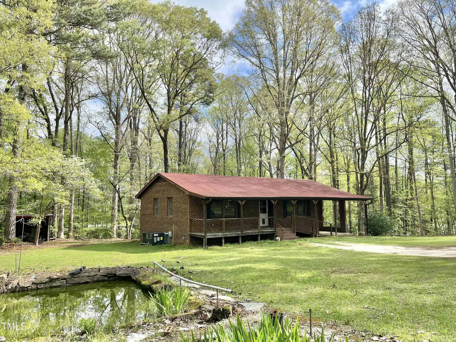 A one-story brick house with a red roof sits in a grassy clearing surrounded by tall trees. A small pond is in the foreground, and a gravel driveway leads up to the house. The sky is partly cloudy.