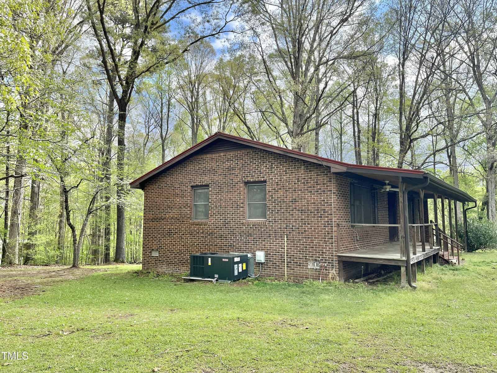 A brick house with a covered porch sits in a wooded area. An air conditioning unit and electric meters are visible along the side. The lawn is grassy, and tall trees surround the house under a partly cloudy sky.
