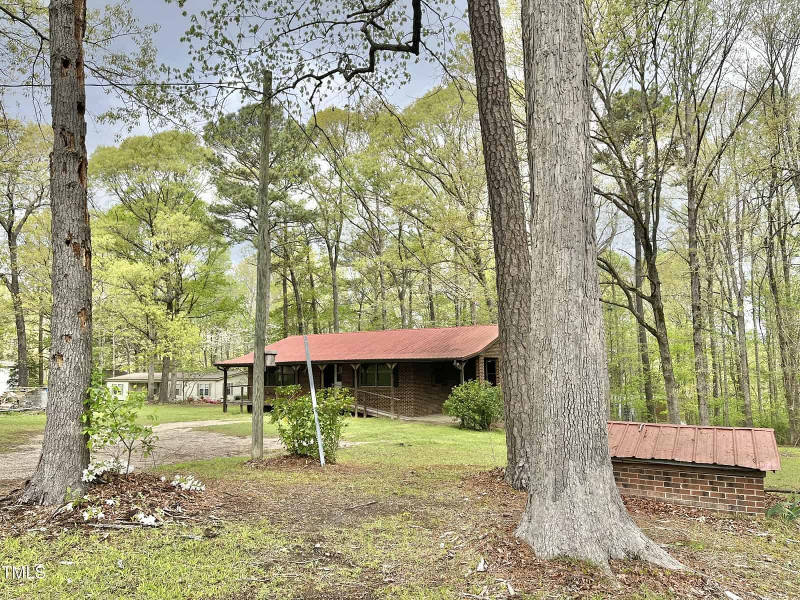 A single-story brick house with a red roof sits among tall trees and green grass. A small brick structure with a matching red roof is in the foreground, and the background is filled with dense leafy woodland.