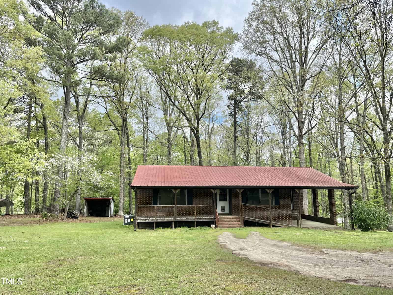 A one-story brick house with a red metal roof sits on a grass lawn surrounded by tall trees. A small covered carport is on the right, and a shed is visible in the background to the left.