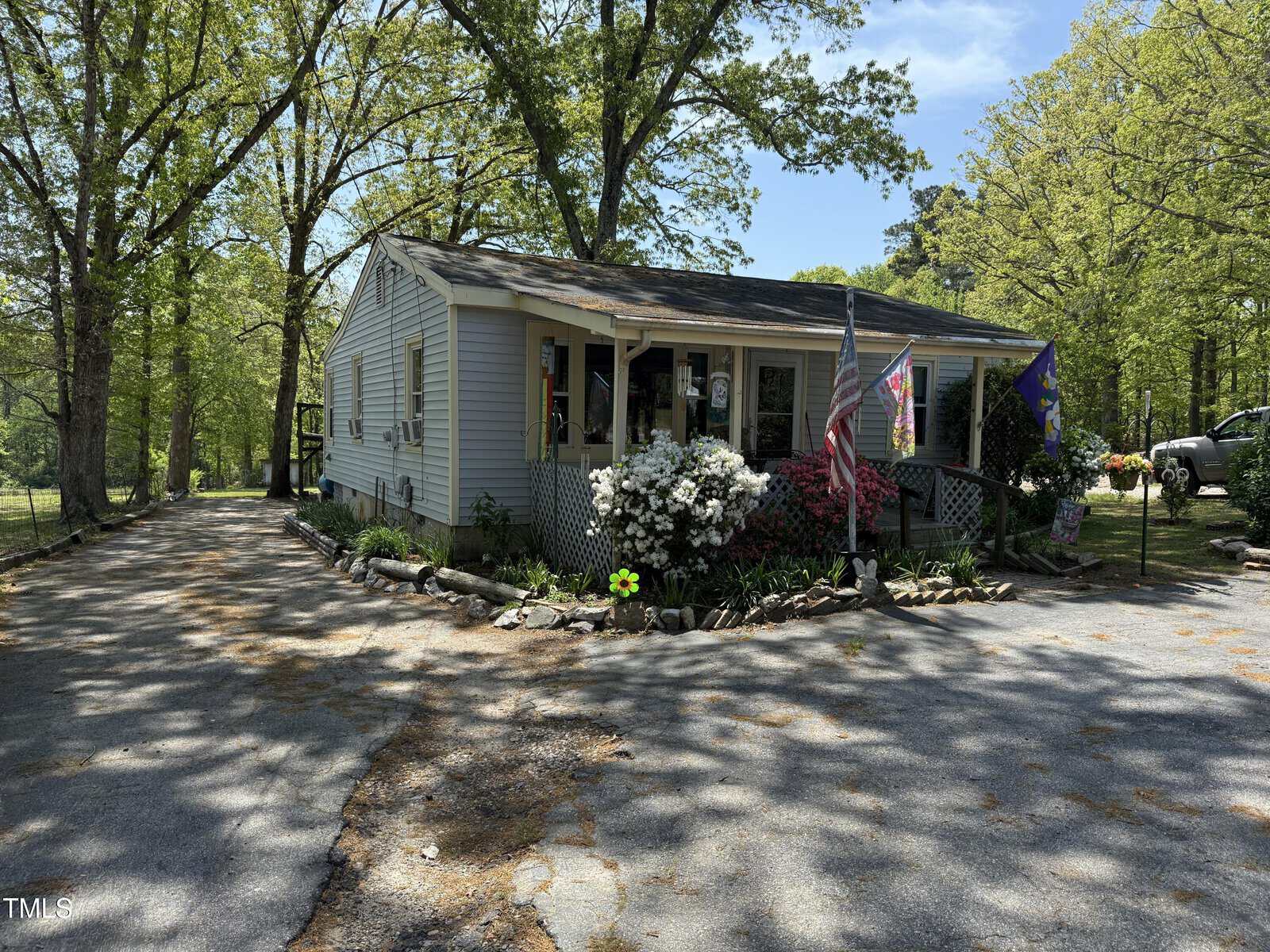 A small single-story house with a sloped roof, front porch, and garden beds along the facade. Two flags are displayed by the entrance. The house is surrounded by trees, with a paved driveway on the left.