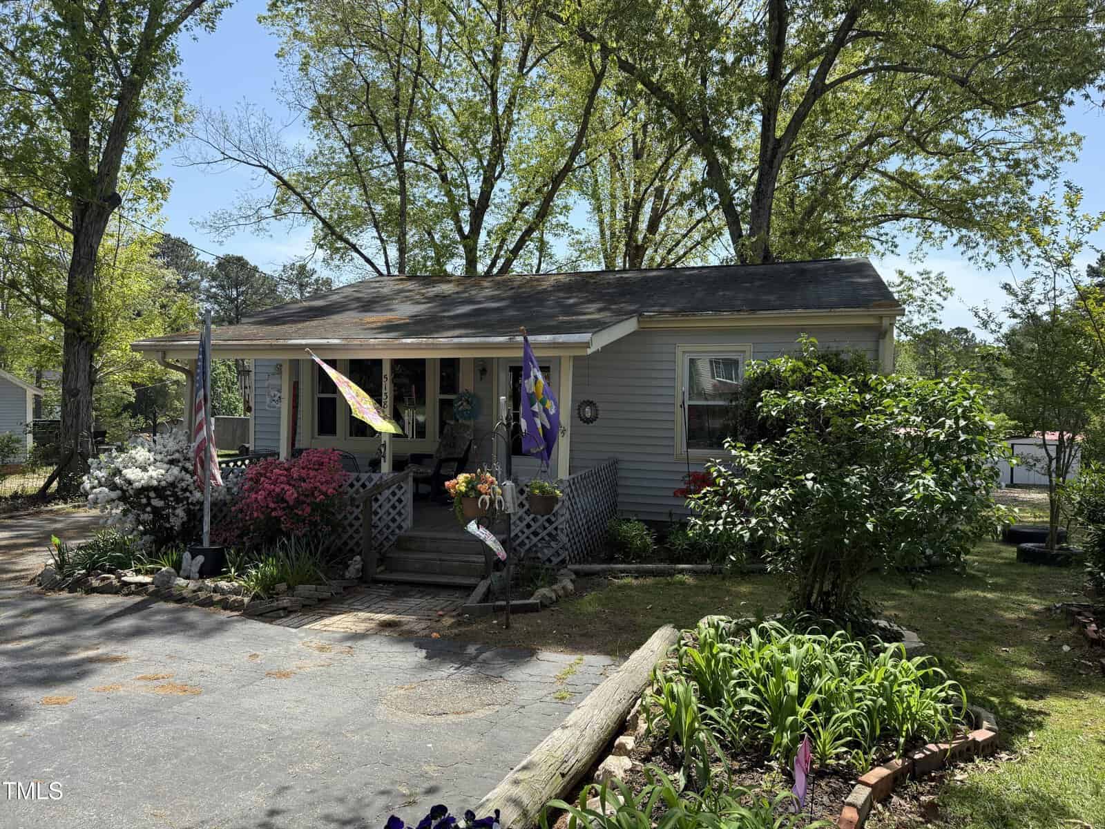 A small gray house with a covered front porch, multiple flags, colorful flowers, and shrubs in the yard. The driveway is paved, and large trees provide shade around the home on a sunny day.