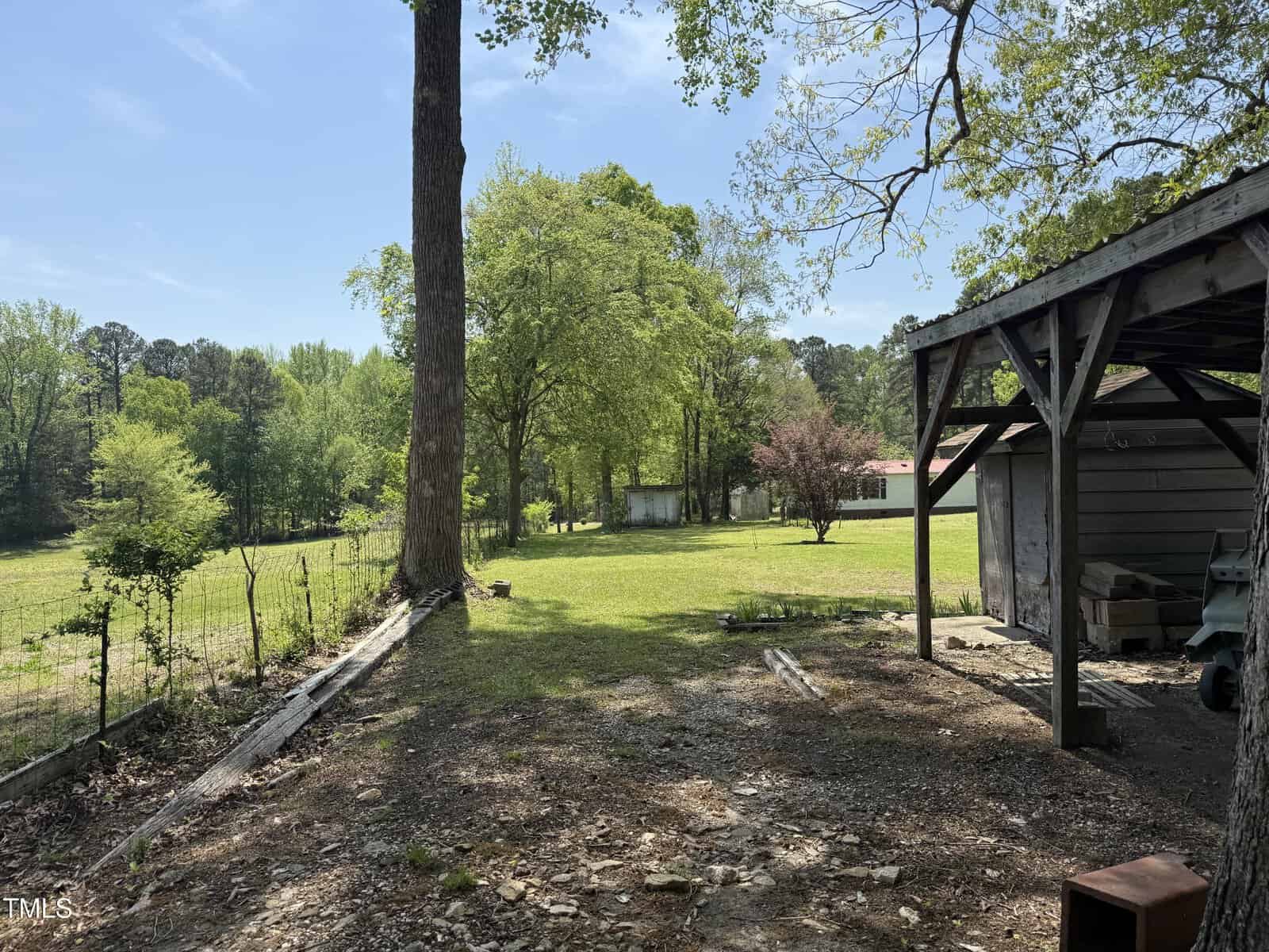 A shaded area with a wooden structure and shed in the foreground opens to a grassy yard, bordered by trees and a wire fence; a metal-roofed building and more trees are visible in the background.